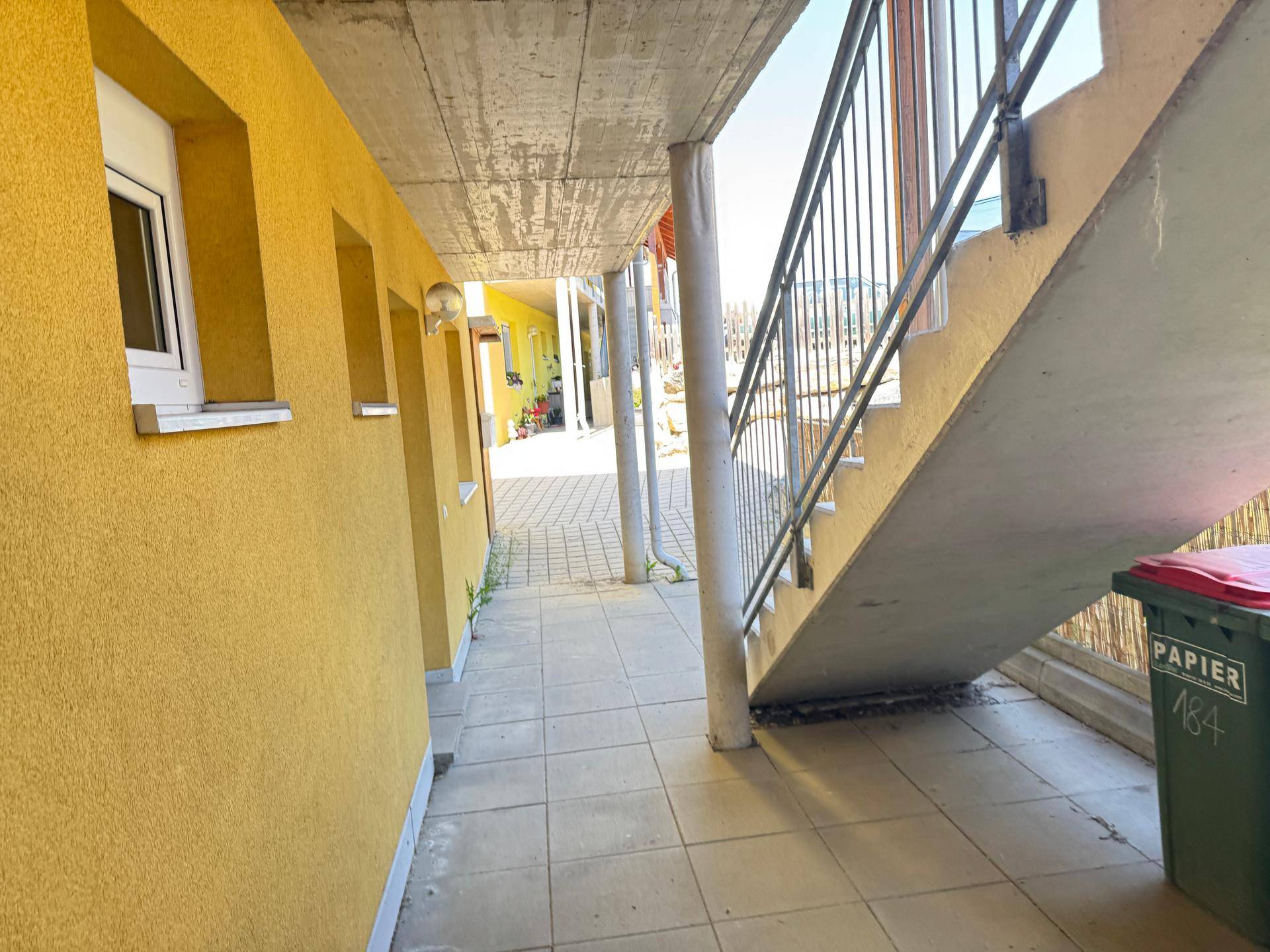Überdachter Gang mit Treppe und gelber Wand, führt zu weiteren Gebäudeteilen.