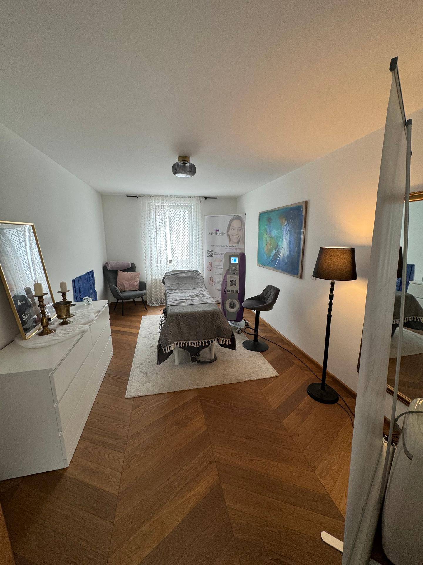 Heller Behandlungsraum mit Liege, Holzparkett und einem großen Spiegel, modern eingerichtet.