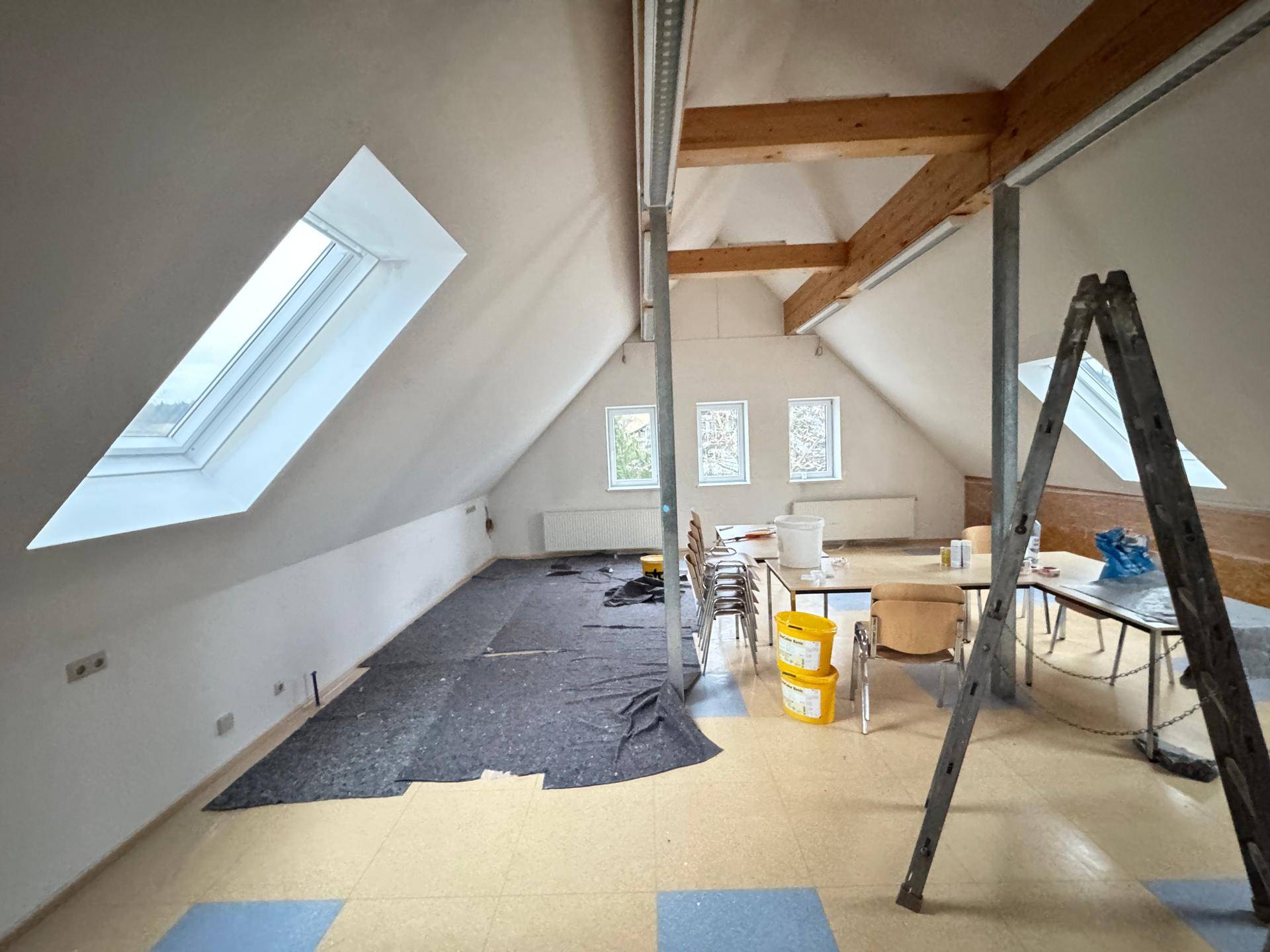 Dachbodenraum mit sichtbaren Holzbalken, Dachfenstern und teilweise abgedecktem Boden.