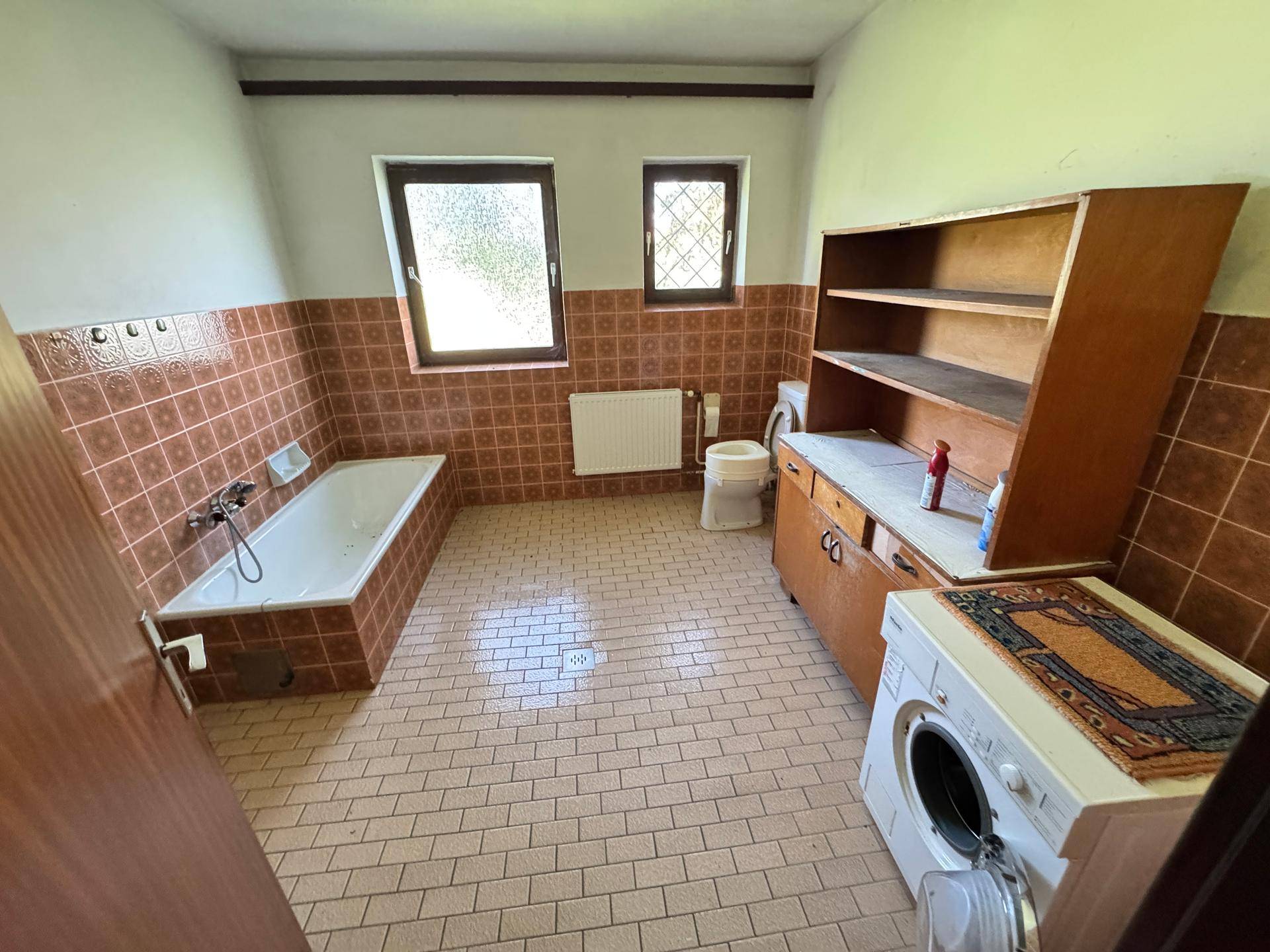 Älteres Badezimmer mit braunen Fliesen, Badewanne, Toilette und Waschmaschine.