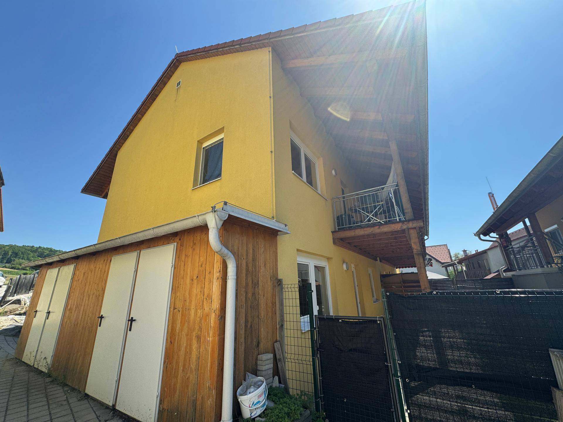 Gelbes Wohngebäude mit Holzbalkon und angrenzenden Abstellräumen unter strahlendem Sonnenschein.