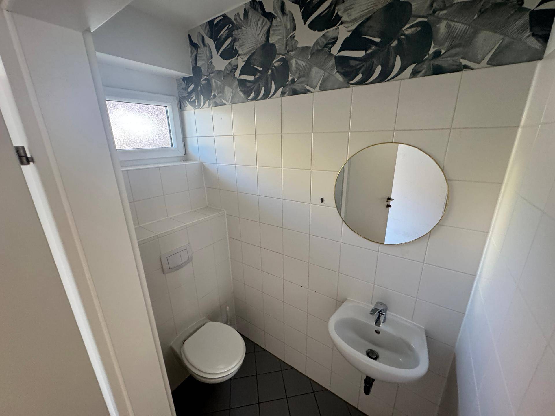 Gäste-WC mit Toilette, kleinem Waschbecken, rundem Spiegel und dekorativer Tapete.