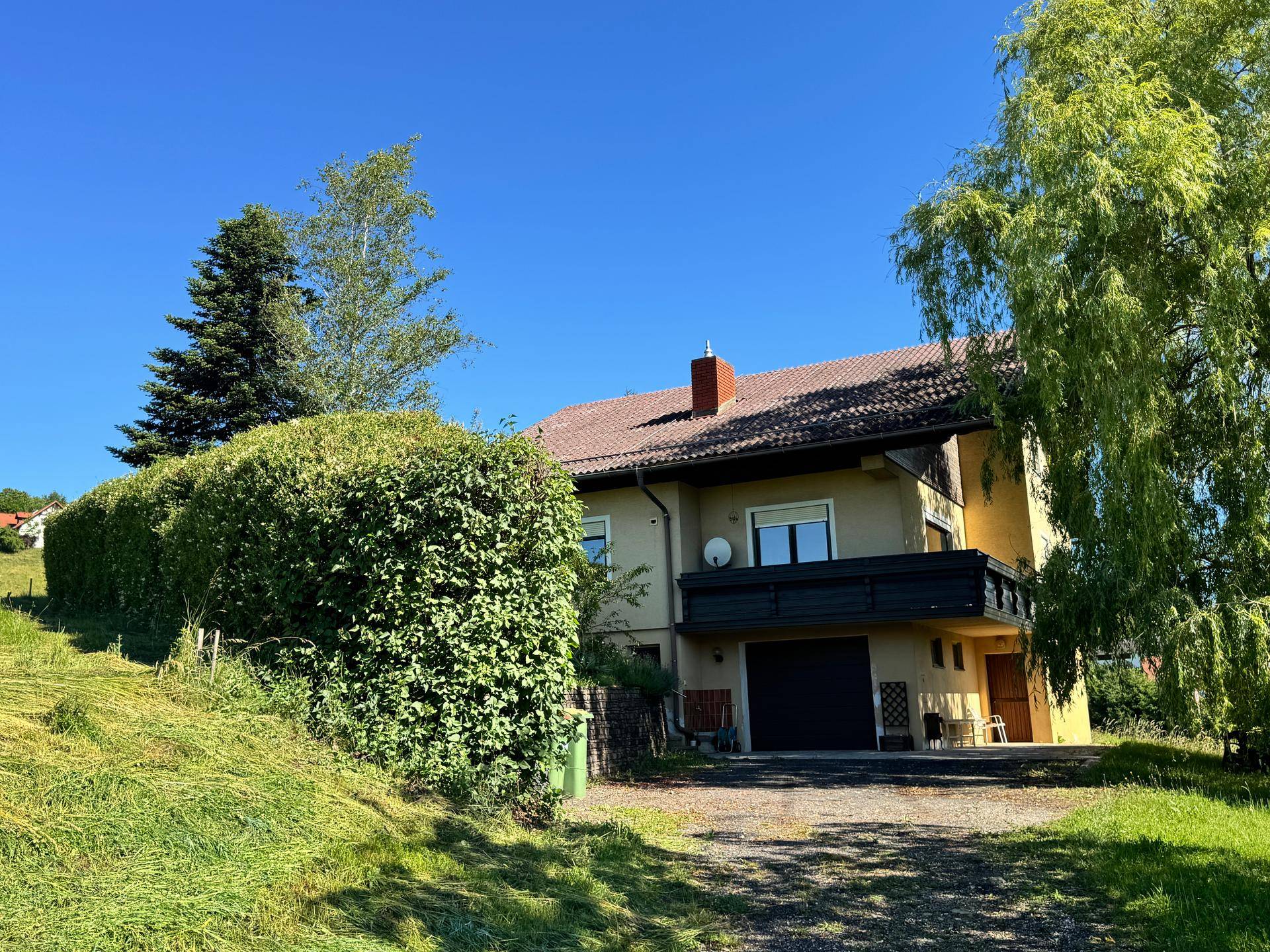 Hausansicht mit Balkon und Garage, umgeben von einer hohen Hecke und Bäumen.