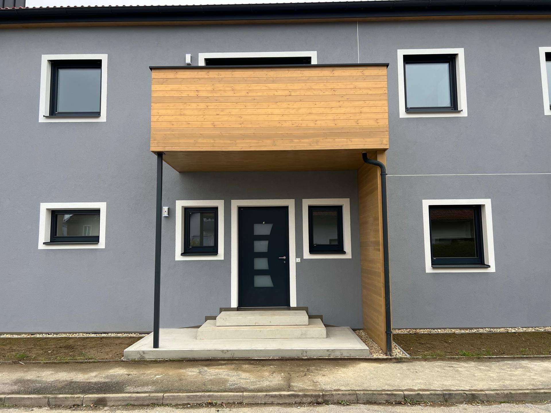 Moderner Eingangsbereich mit dunkler Tür, grauer Fassade und dekorativen Holzelementen.