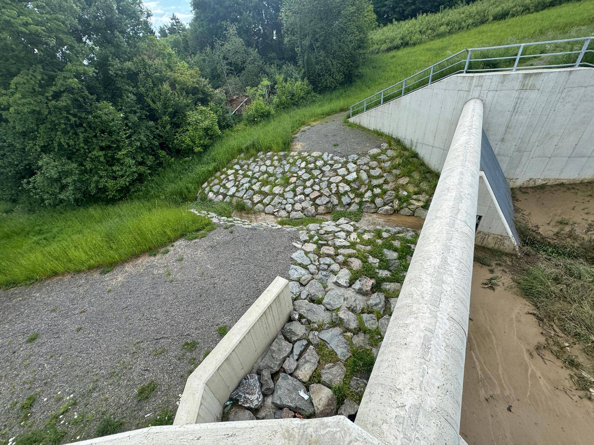 Betonierte Entwässerungsanlage mit Steinen und grünen Hängen in einer ländlichen Umgebung.