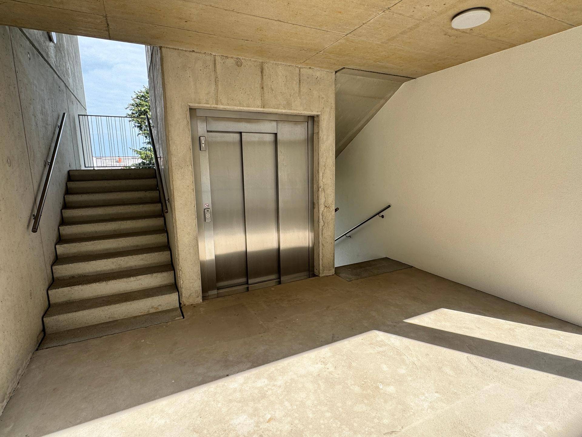 Moderner Aufzug und Treppenhaus in einem Gebäude mit Sichtbetonwänden und klaren Linien.