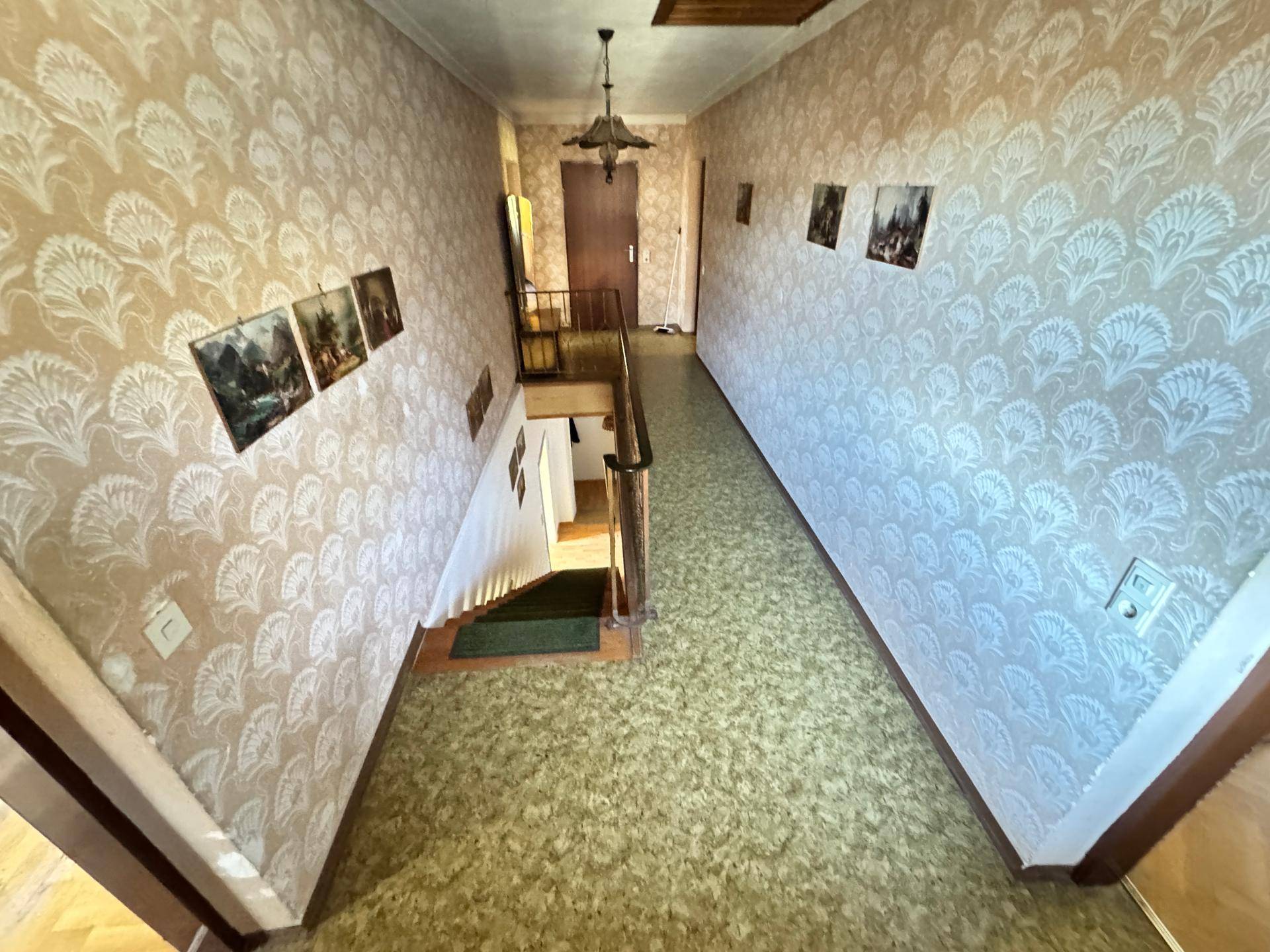 Treppenhaus und Flur mit gemusterter Tapete, Teppichboden und einem schmiedeeisernen Geländer.