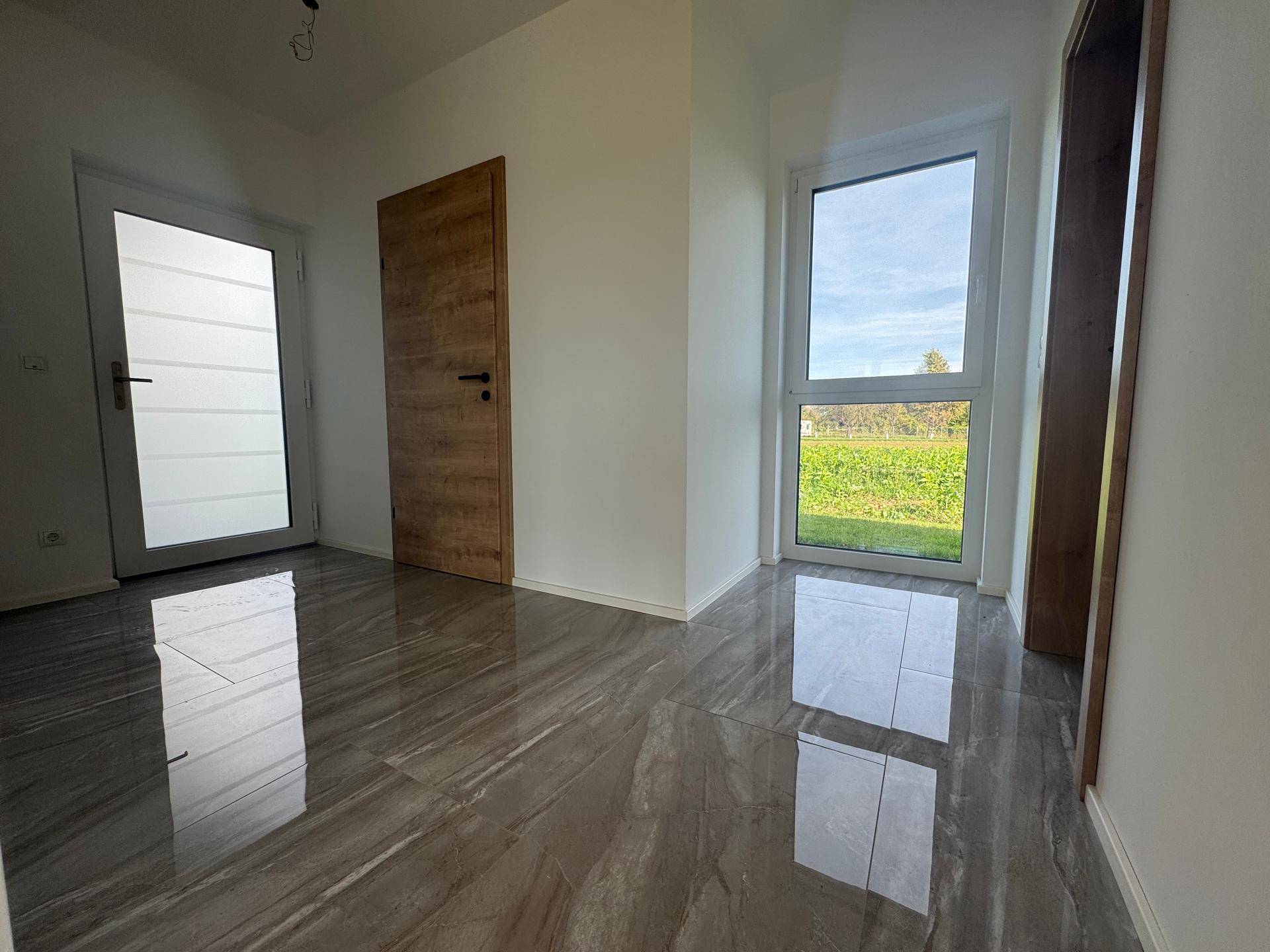 Lichtdurchfluteter Eingangsbereich mit grauen Fliesen, Holztür und Blick ins Grüne.