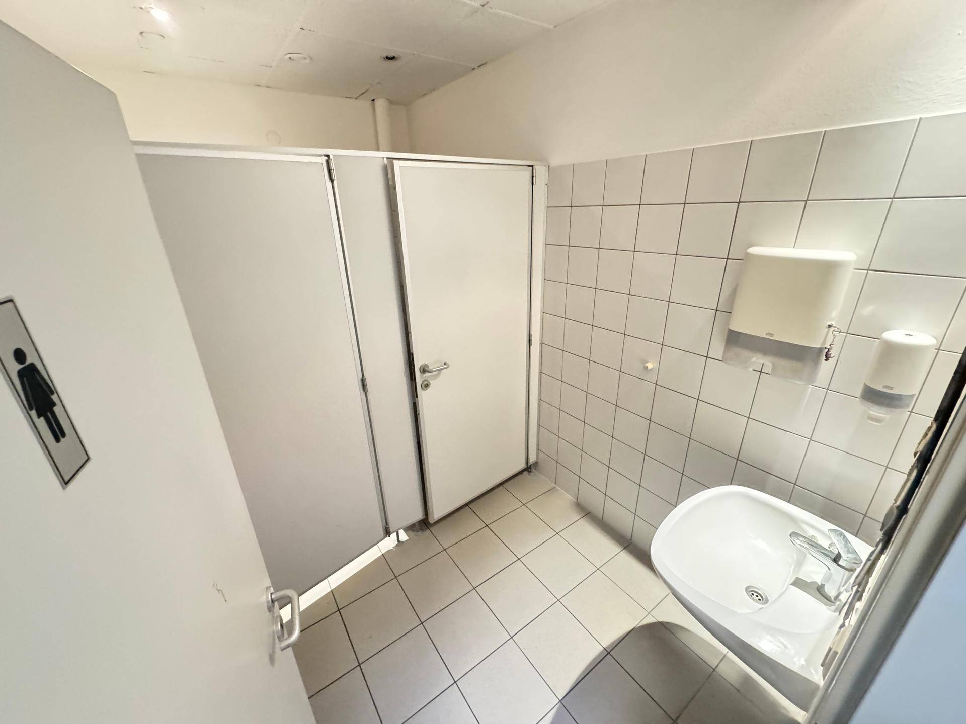 Öffentlicher Toilettenbereich mit mehreren Kabinen, Waschbecken und weiß gefliesten Wänden.