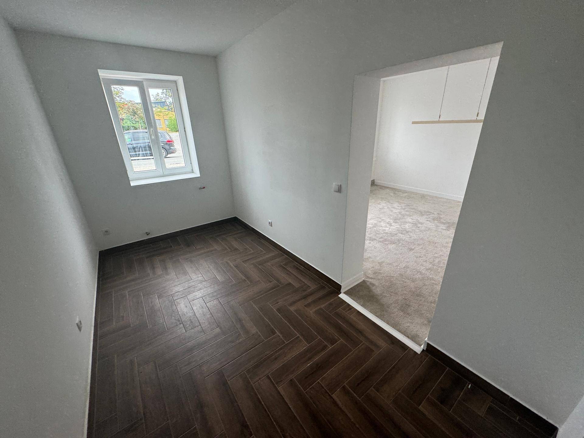 Zimmer mit dunklem Fischgrätparkett und offenem Zugang zu einem weiteren, hellen Raum.