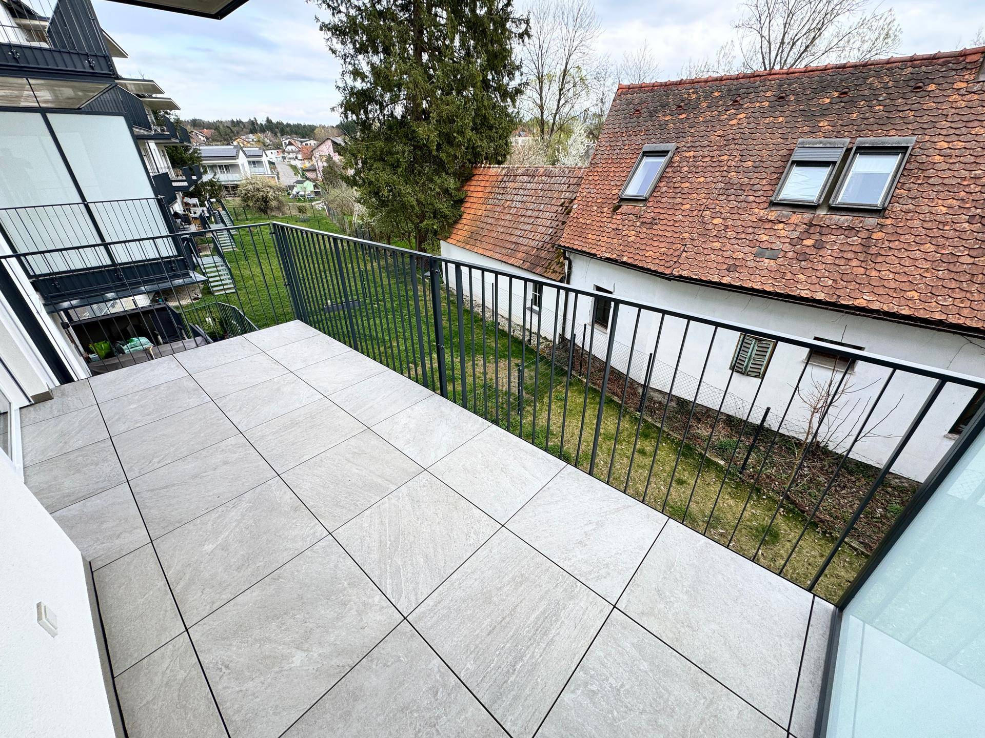 Geräumiger Balkon mit Fliesenboden und schwarzem Geländer, bietet einen weiten Blick ins Grüne.