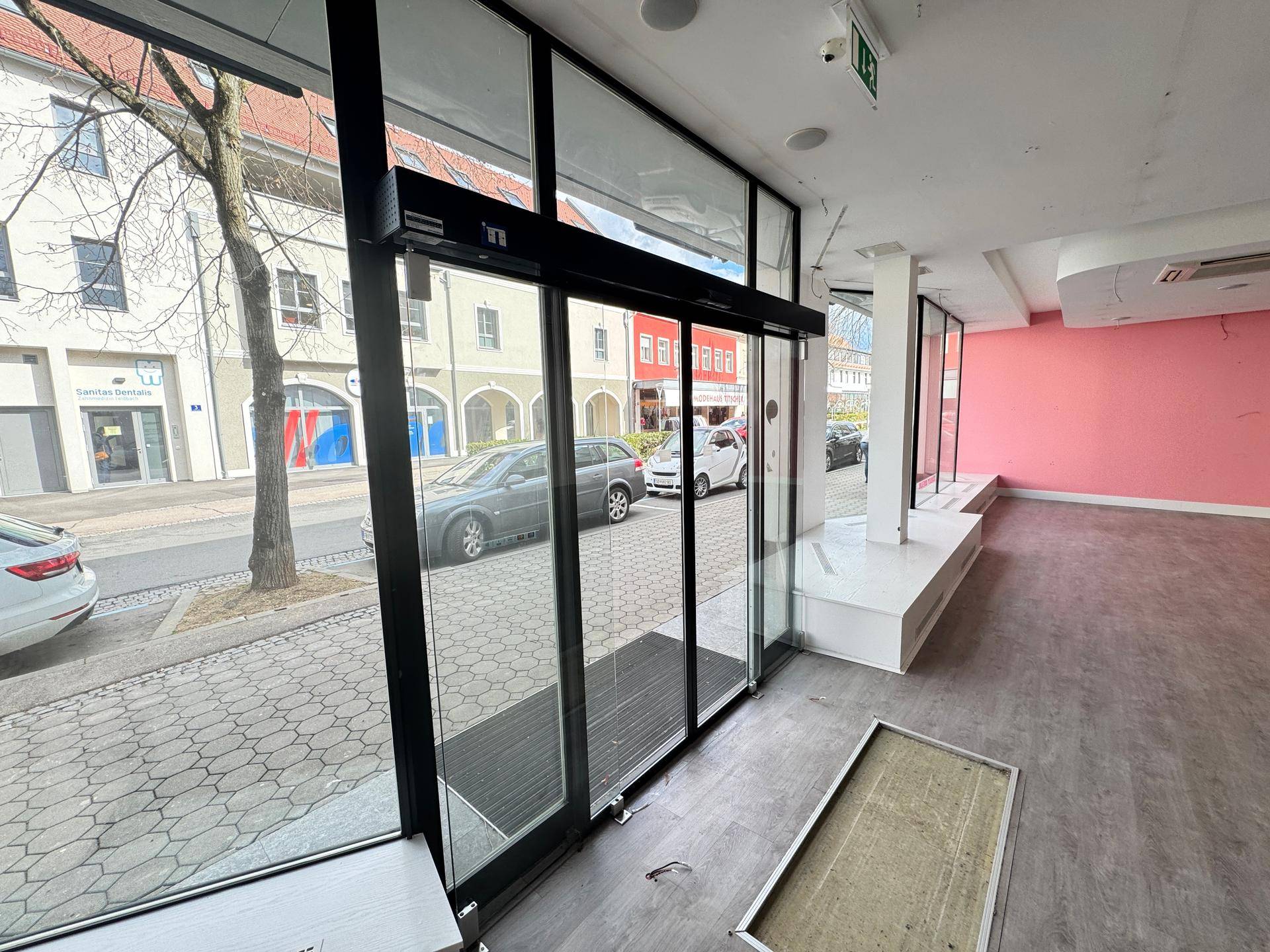 Innenansicht eines leeren Ladenlokals mit großen Schaufenstern und rosa Wandakzenten.