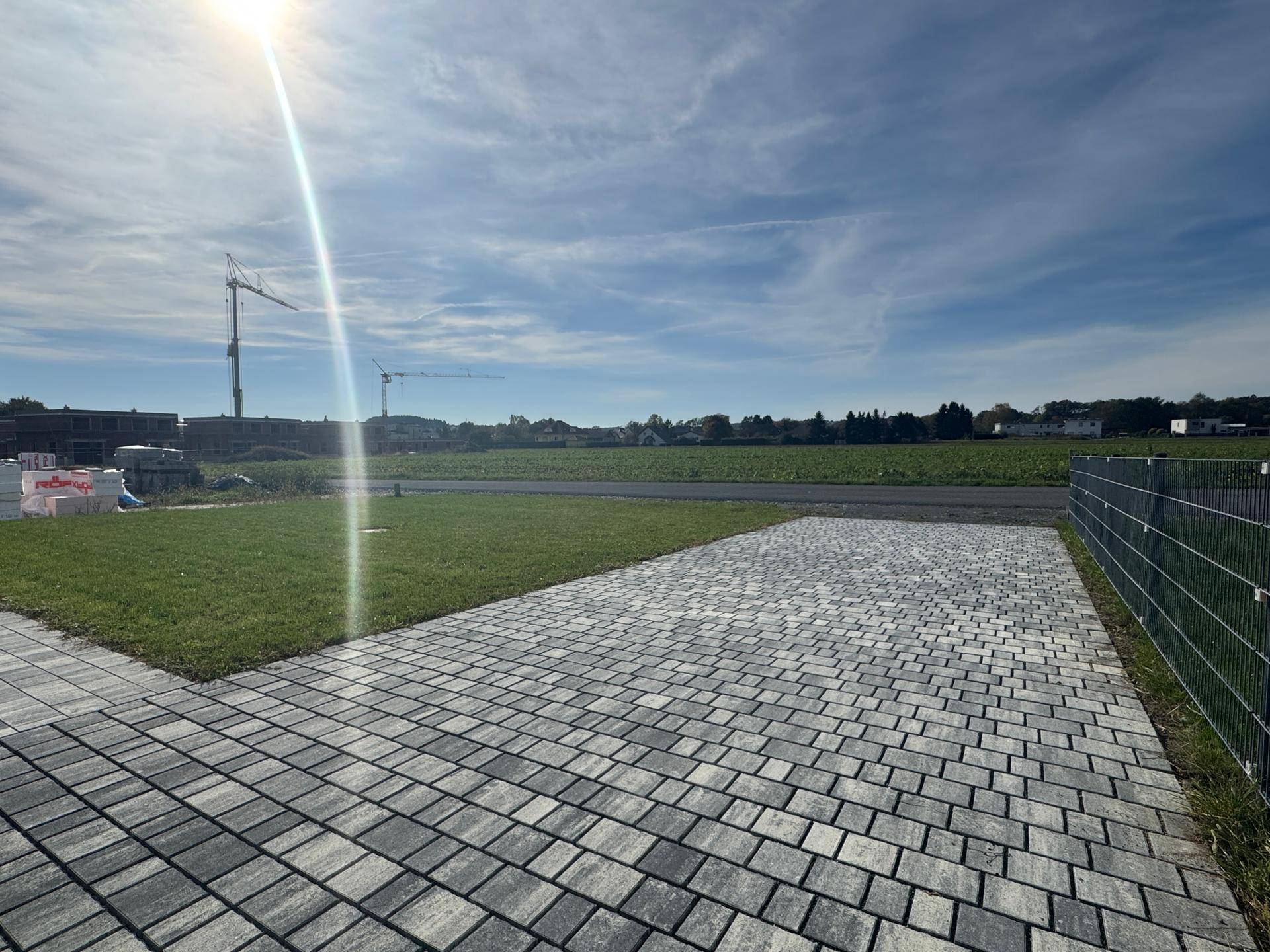 Gepflasterter Außenbereich neben Rasenfläche mit Blick auf Felder und Baustellenkräne.