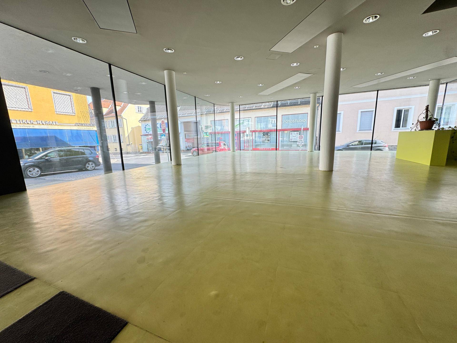 Heller, offener Raum mit großen Fensterfronten und grünem Bodenbelag, ideal für flexible Nutzung und viel Tageslicht.