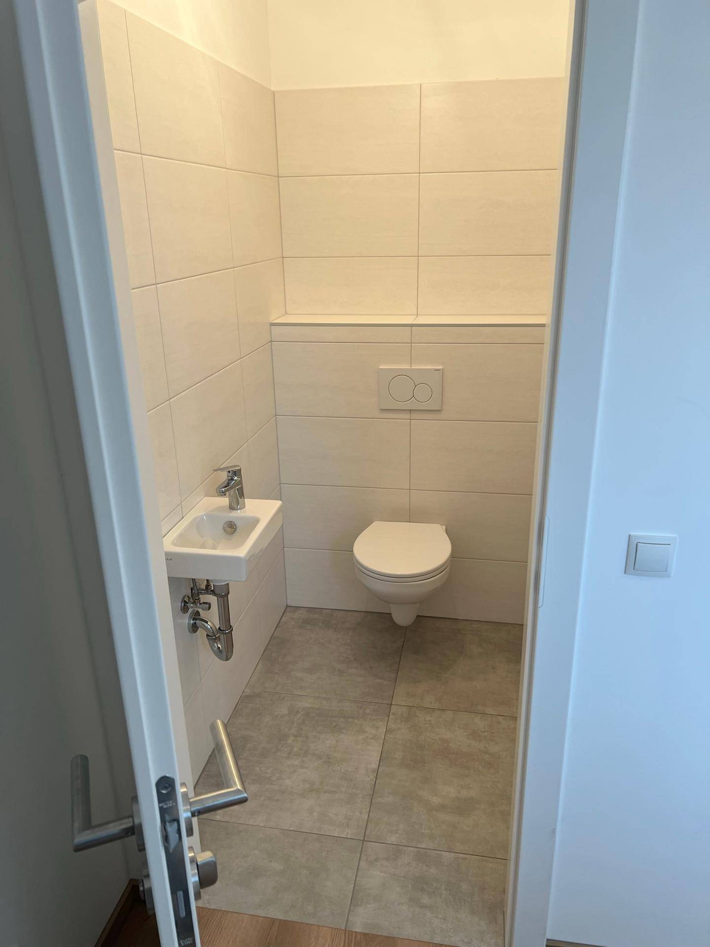 Modernes Gäste-WC mit wandhängendem WC, kleinem Waschbecken und hellen Fliesen an den Wänden.