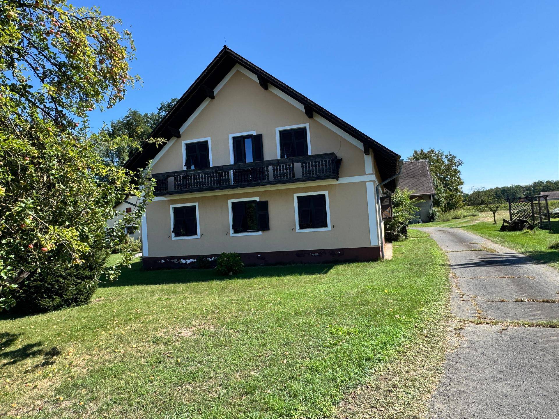 Haus mit Balkon, grüner Wiese und Zufahrtsweg in ländlicher Umgebung unter blauem Himmel.