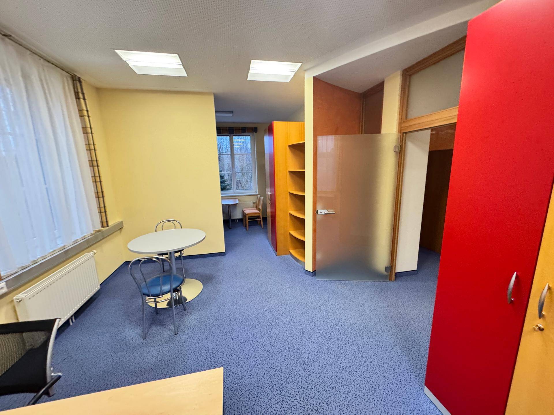 Büroraum mit bunten Schränken, blauem Teppichboden und einem kleinen Besprechungstisch.
