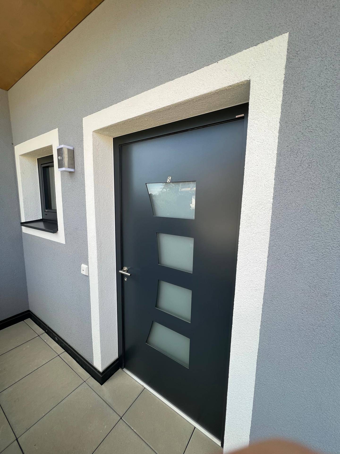 Moderner Eingangsbereich mit dunkler Tür, grauer Fassade und dekorativen Holzelementen.