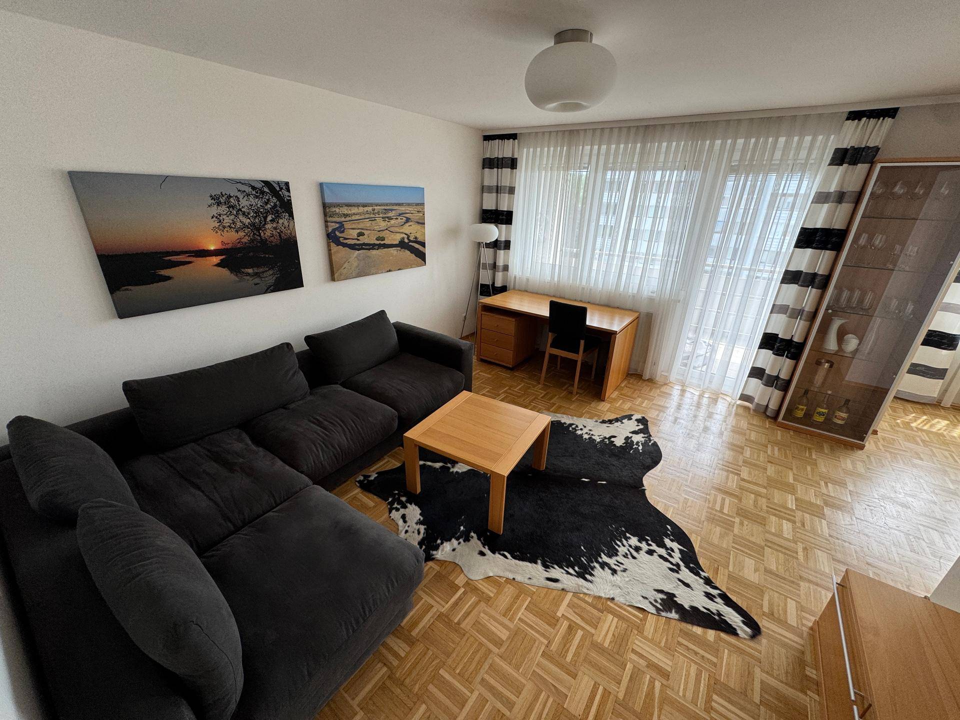 Helles Wohnzimmer mit dunkler Eckcouch, Holztisch und Arbeitsplatz am Fenster.