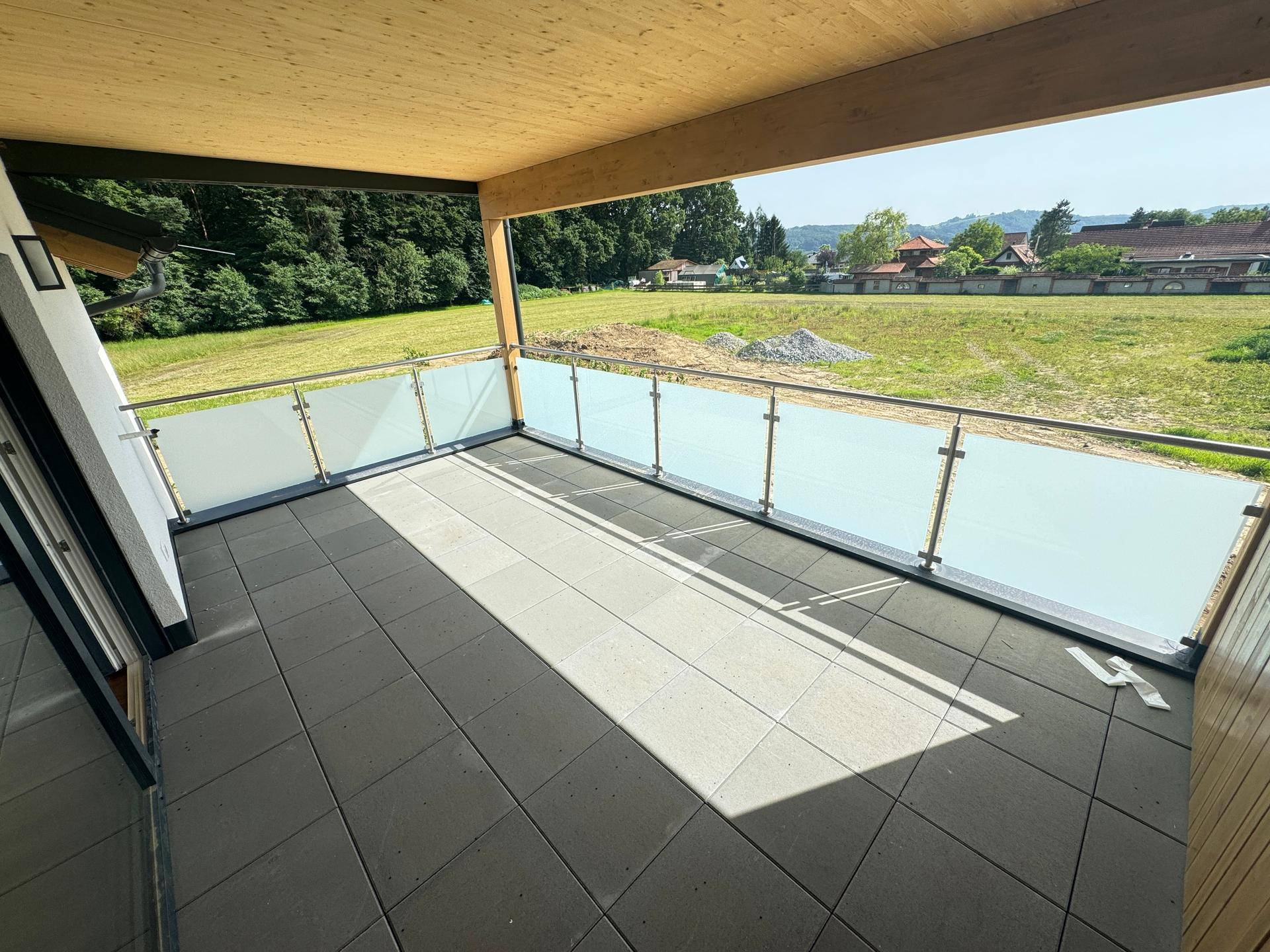 Heller Balkon mit Fliesenboden, Glasgeländer und sonnigem Ausblick in die Natur.