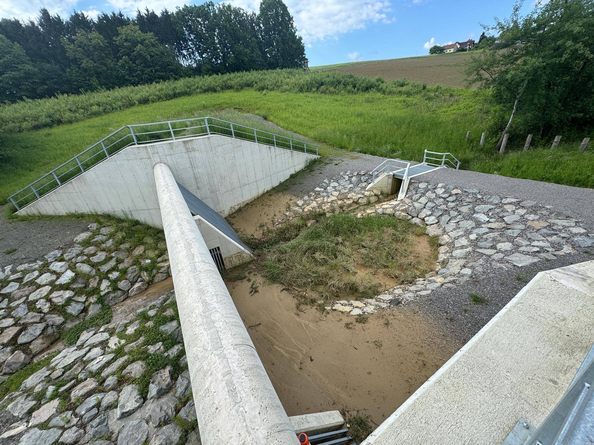 Betonierte Entwässerungsanlage mit Steinen und grünen Hängen, teilweise mit Schlamm gefüllt.