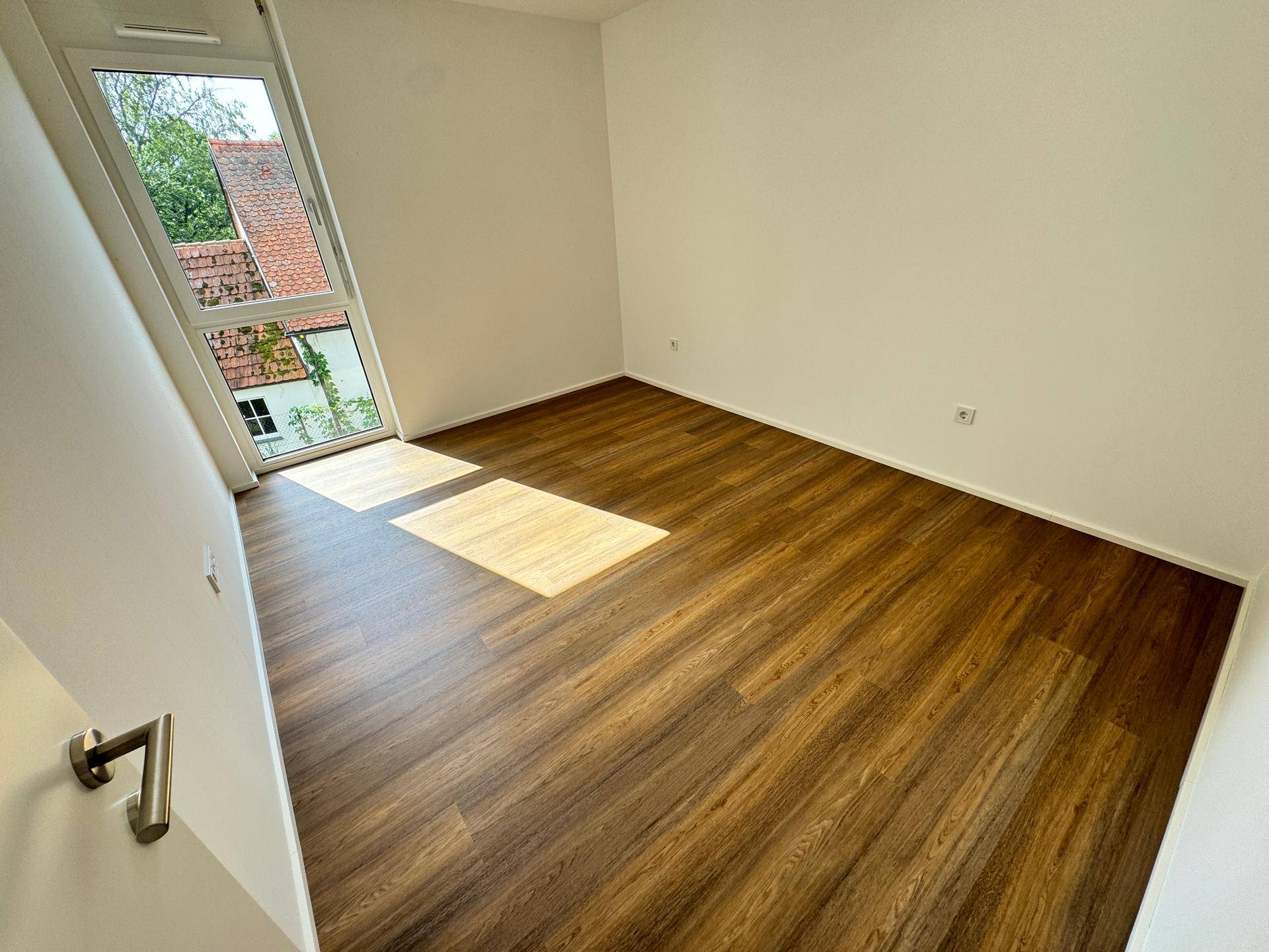 Helles Schlafzimmer mit Holzboden und großem Fenster, das viel Tageslicht hereinlässt.