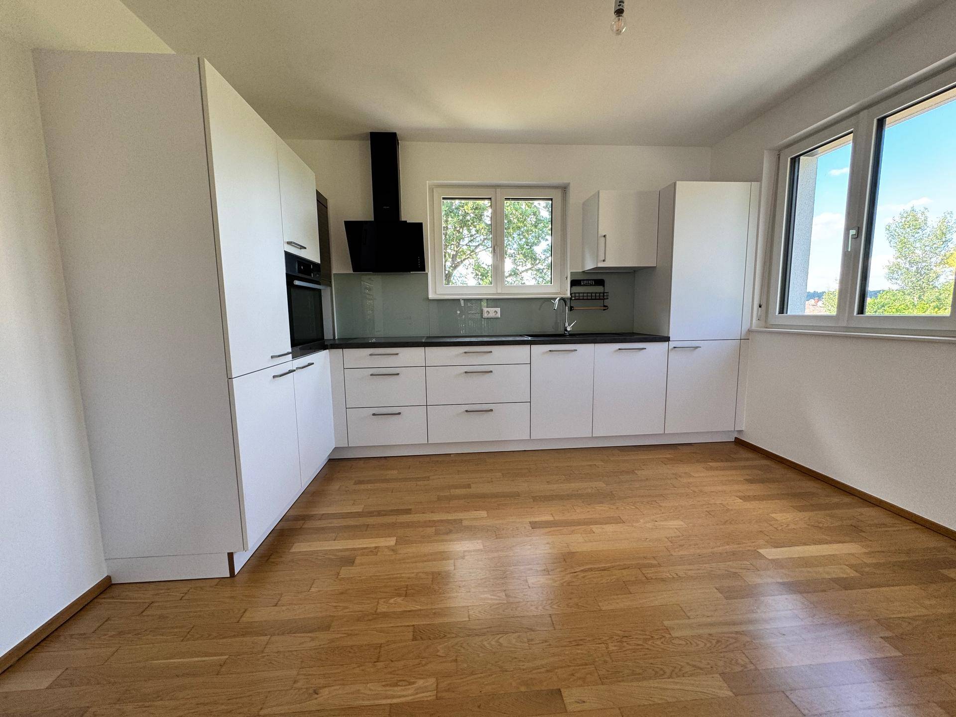Neue, weiße Einbauküche mit dunkler Arbeitsplatte und Holzboden in einem lichtdurchfluteten Raum.