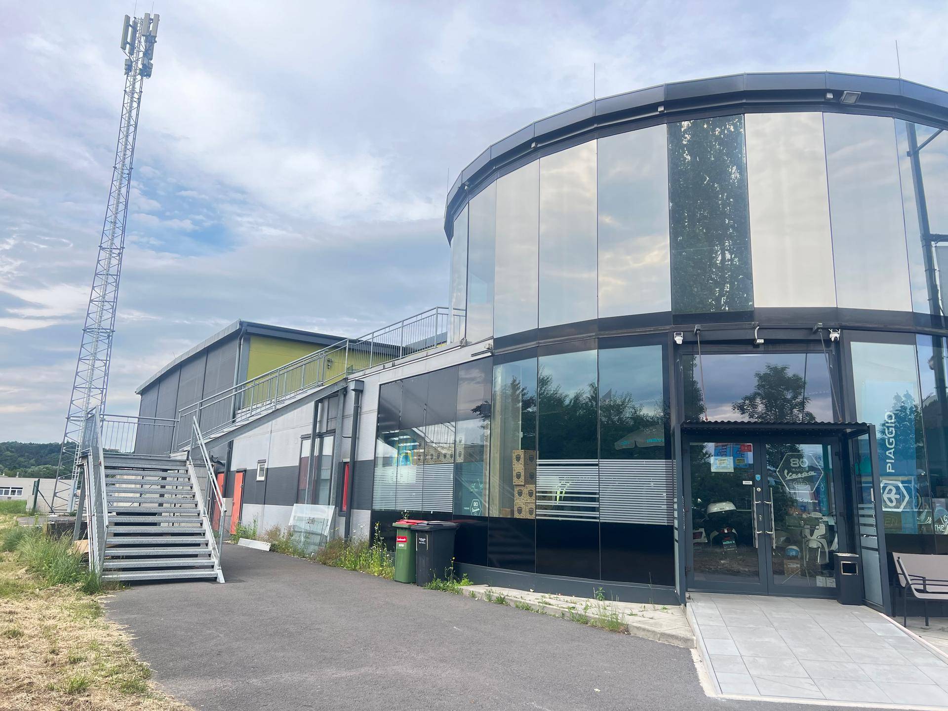 Modernes Gebäude mit runder Glasfassade, Außentreppe und einem Mobilfunkmast im Hintergrund.