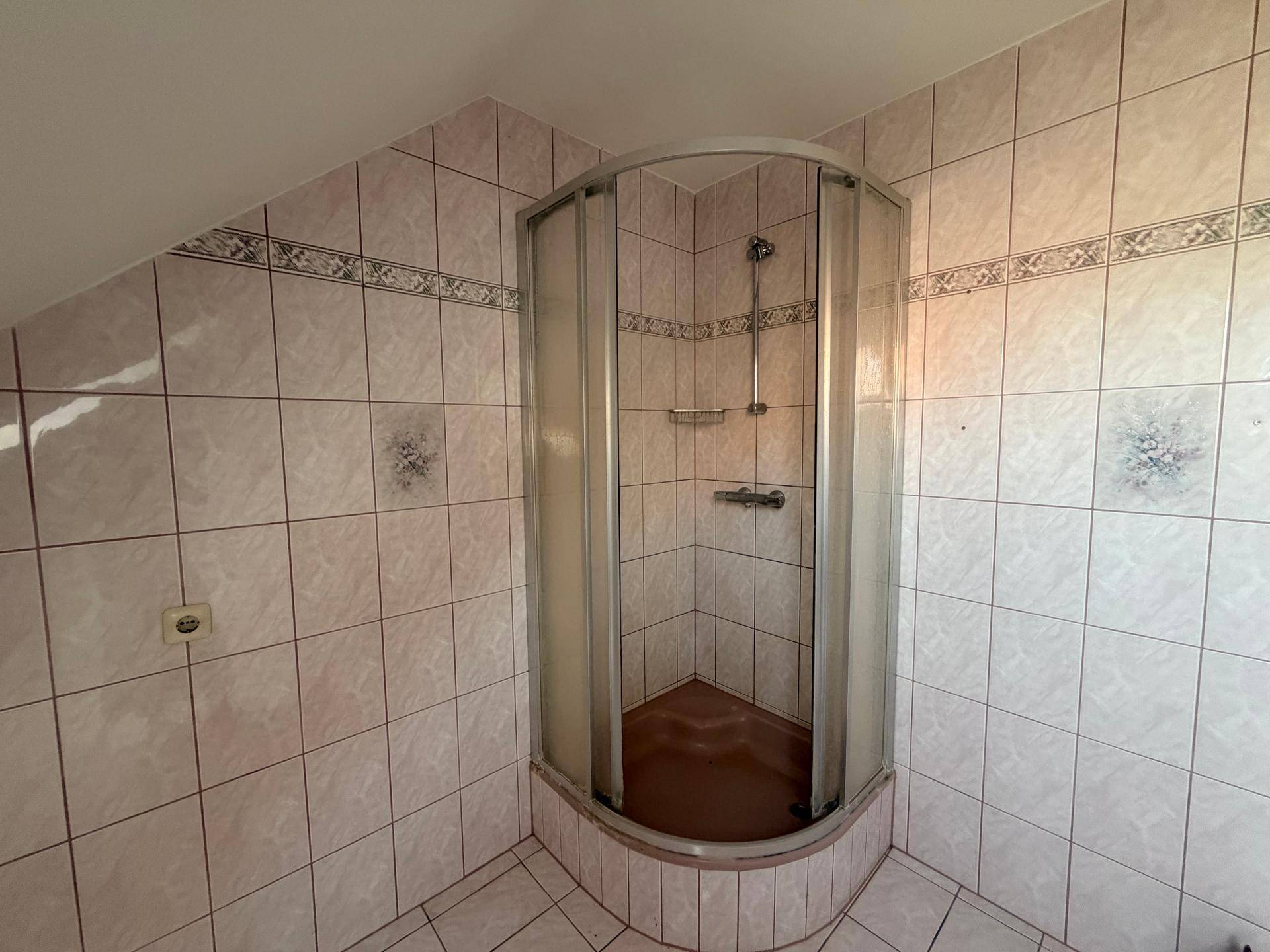 Badezimmer mit Duschkabine, rosa Fliesen und Dachschräge in älterem Zustand.