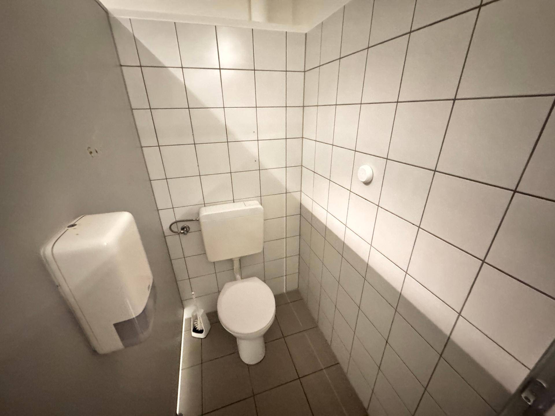 Sauberes WC mit weiß gefliesten Wänden und Boden, ausgestattet mit einer Toilette und Spender.