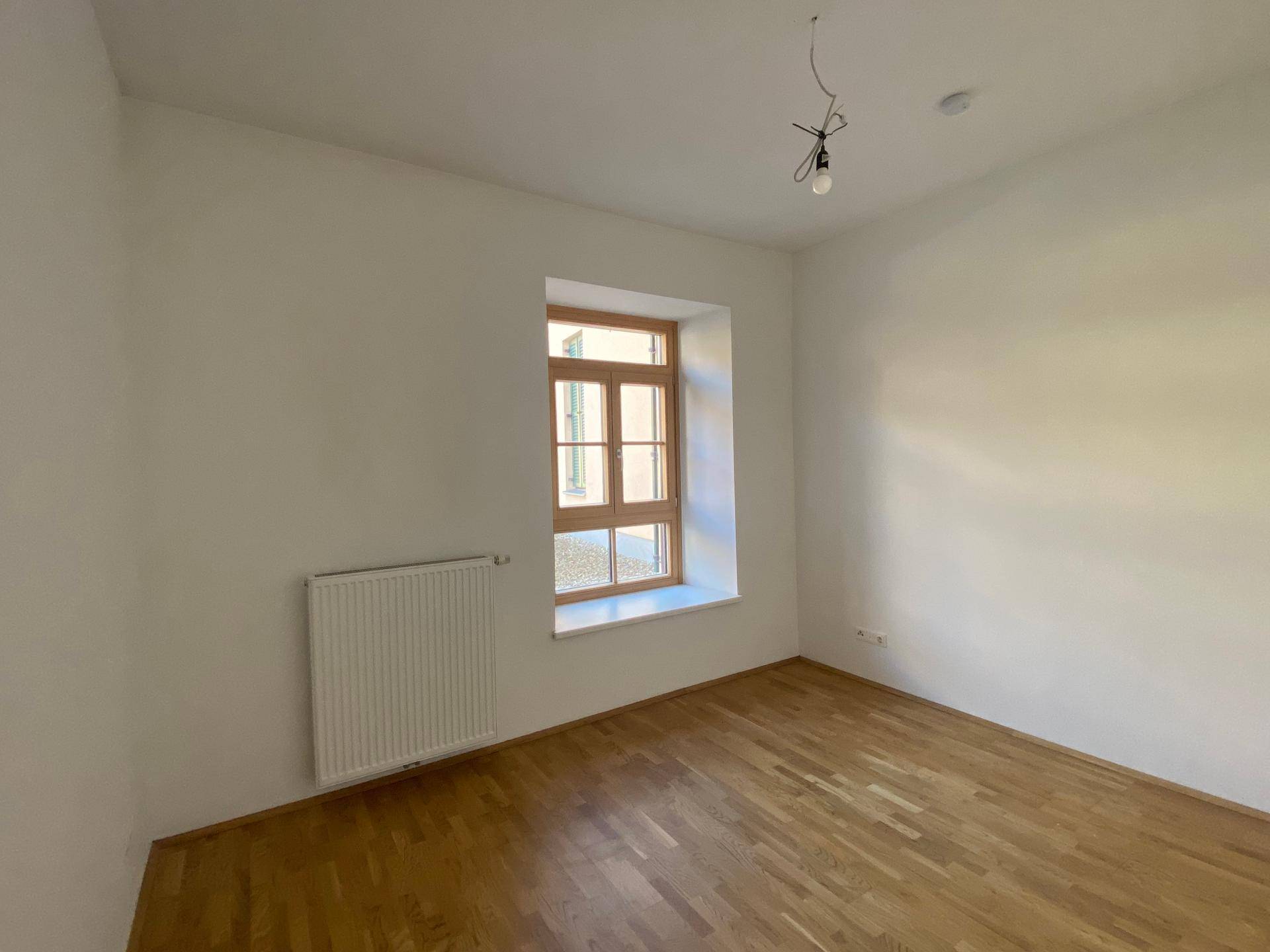 Ruhiges Zimmer mit Parkettboden, weiß gestrichenen Wänden und einem Fenster mit Heizkörper.