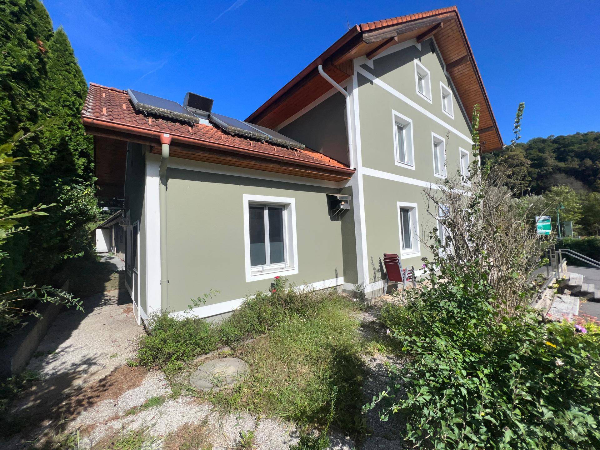 Seitenansicht des Gebäudes mit Solarpaneelen auf dem Dach und umgebender Vegetation.