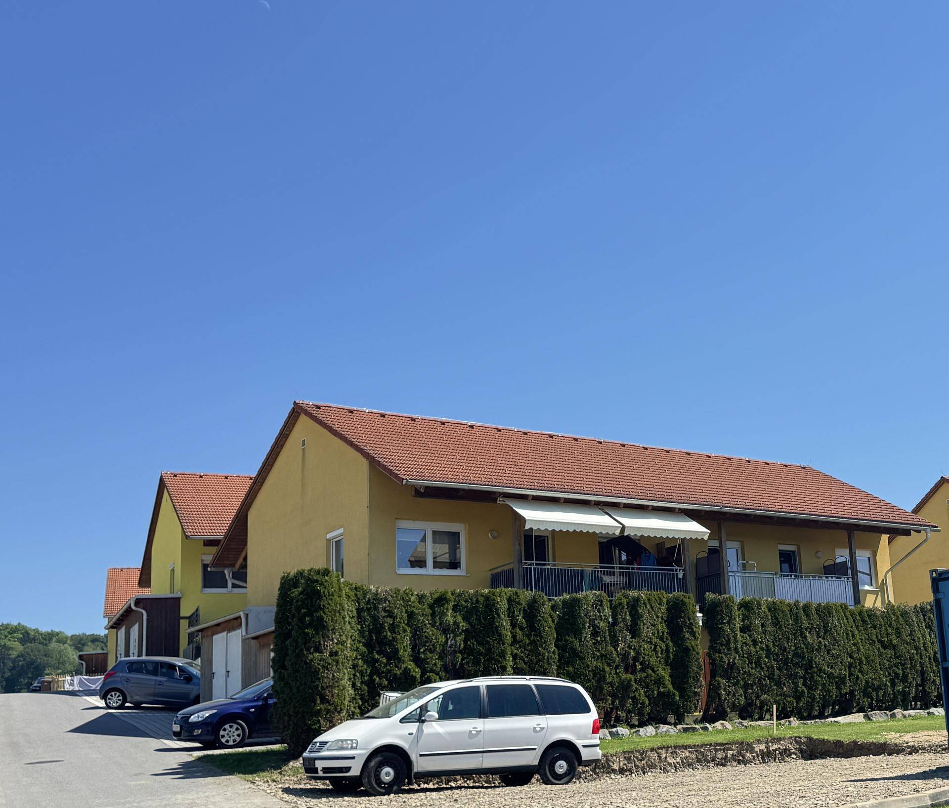 Wohngebäude mit gepflegter Hecke und geparkten Autos auf einem Schotterparkplatz.