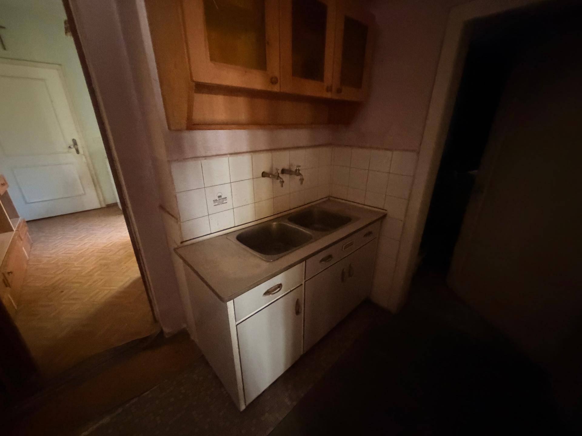 Kleine, einfache Küchenzeile mit Doppelspüle und Hängeschränken in dunklem Raum.