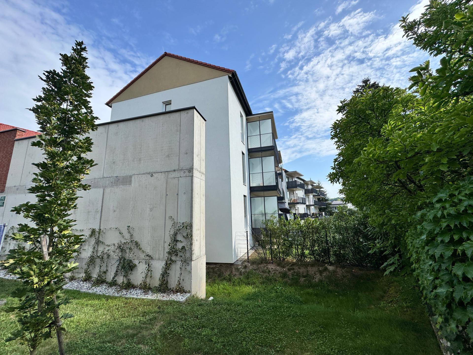 Außenansicht des Neubaus mit Betonwand, Balkonen und umgebenden Grünflächen unter blauem Himmel.