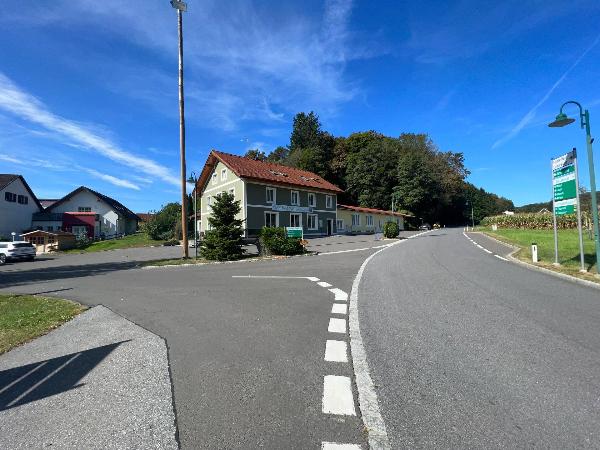 Panoramablick auf das Gebäude mit Maibaum, umgeben von ländlicher Umgebung und einer Straße.
