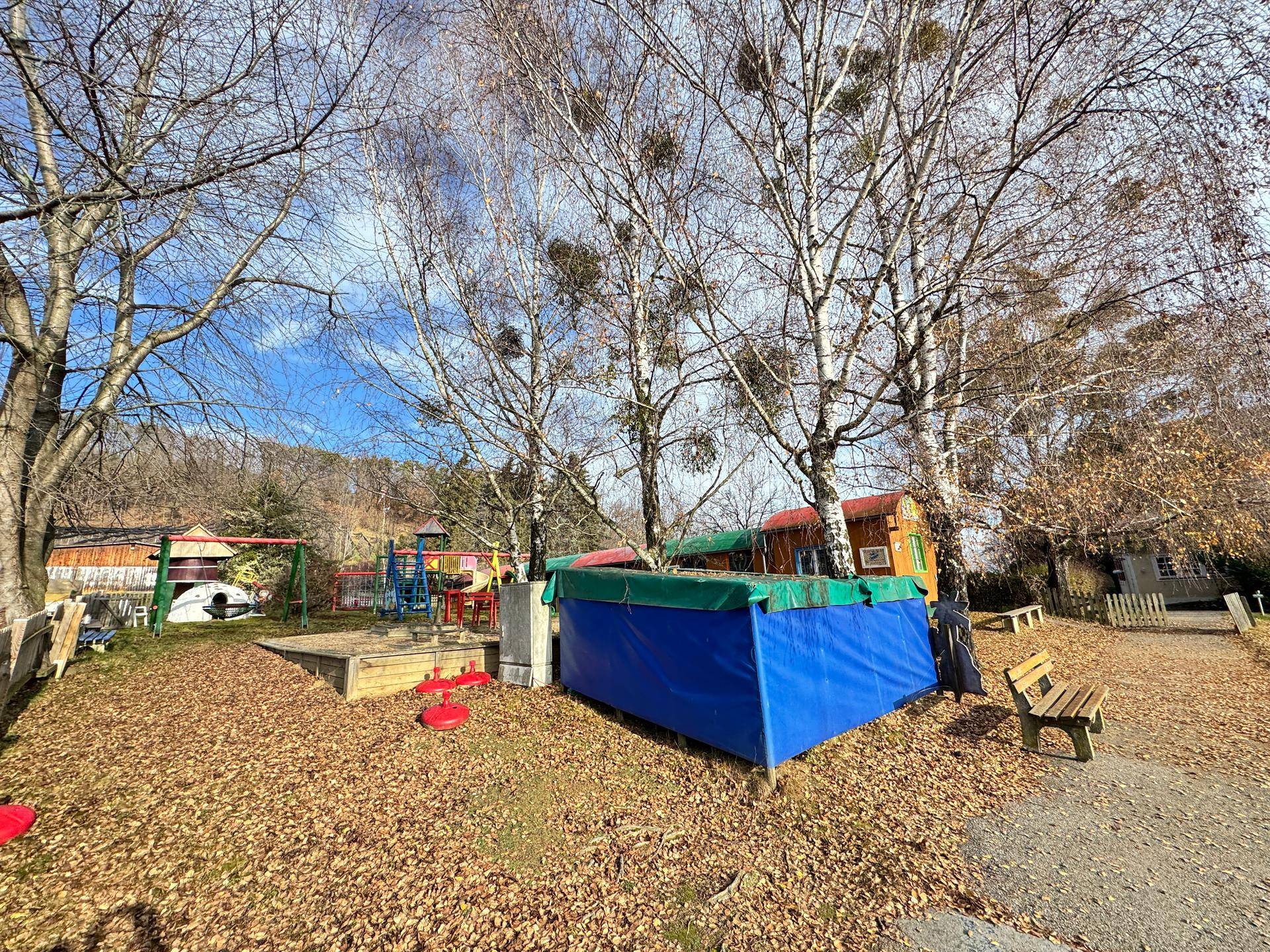 Spielplatz mit Sandkasten, Schaukeln und Bäumen in einem Gartenbereich.