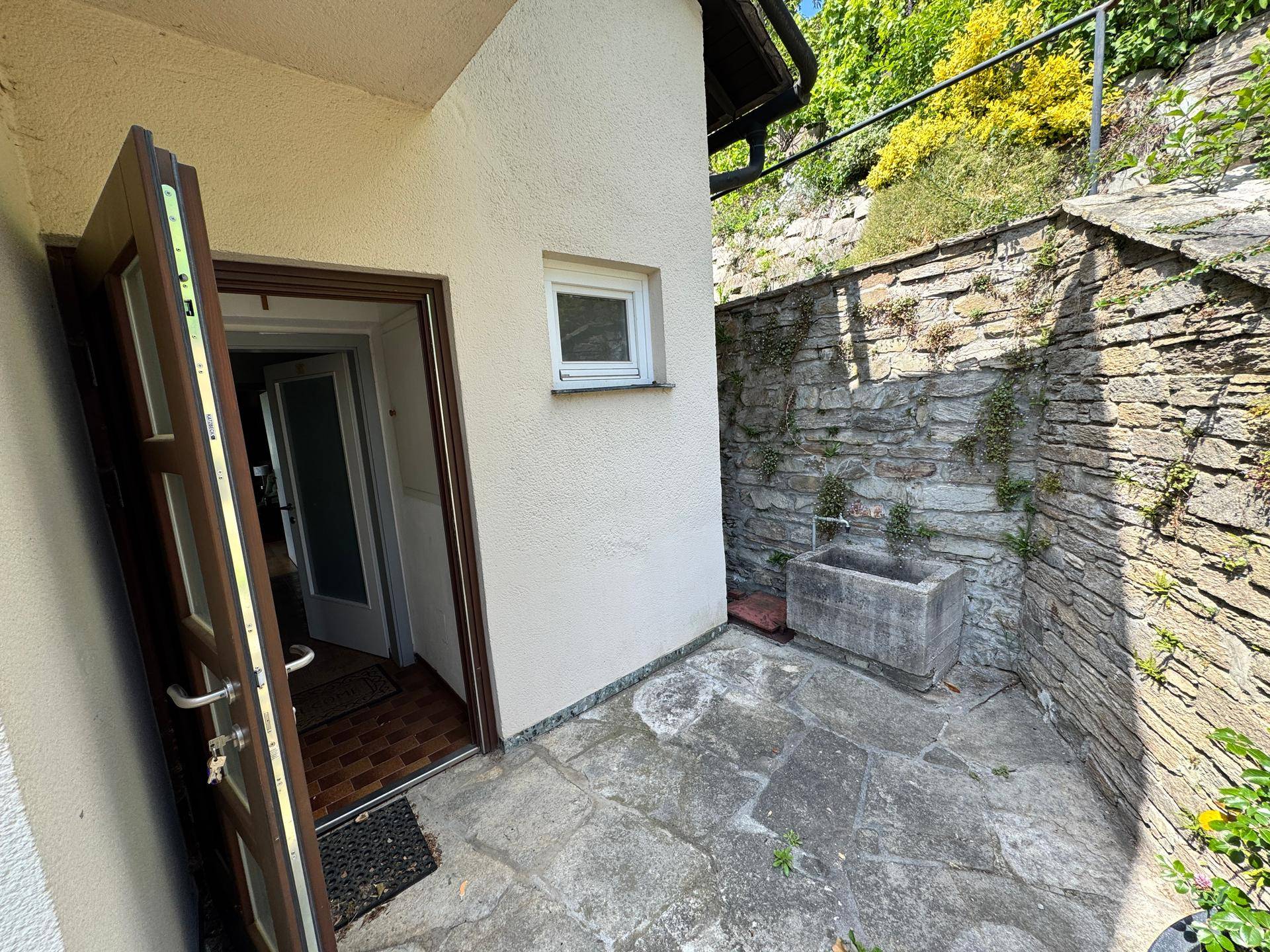 Eingangsbereich mit offener Tür, gepflastertem Patio und rustikaler Steinmauer, umgeben von Grün.