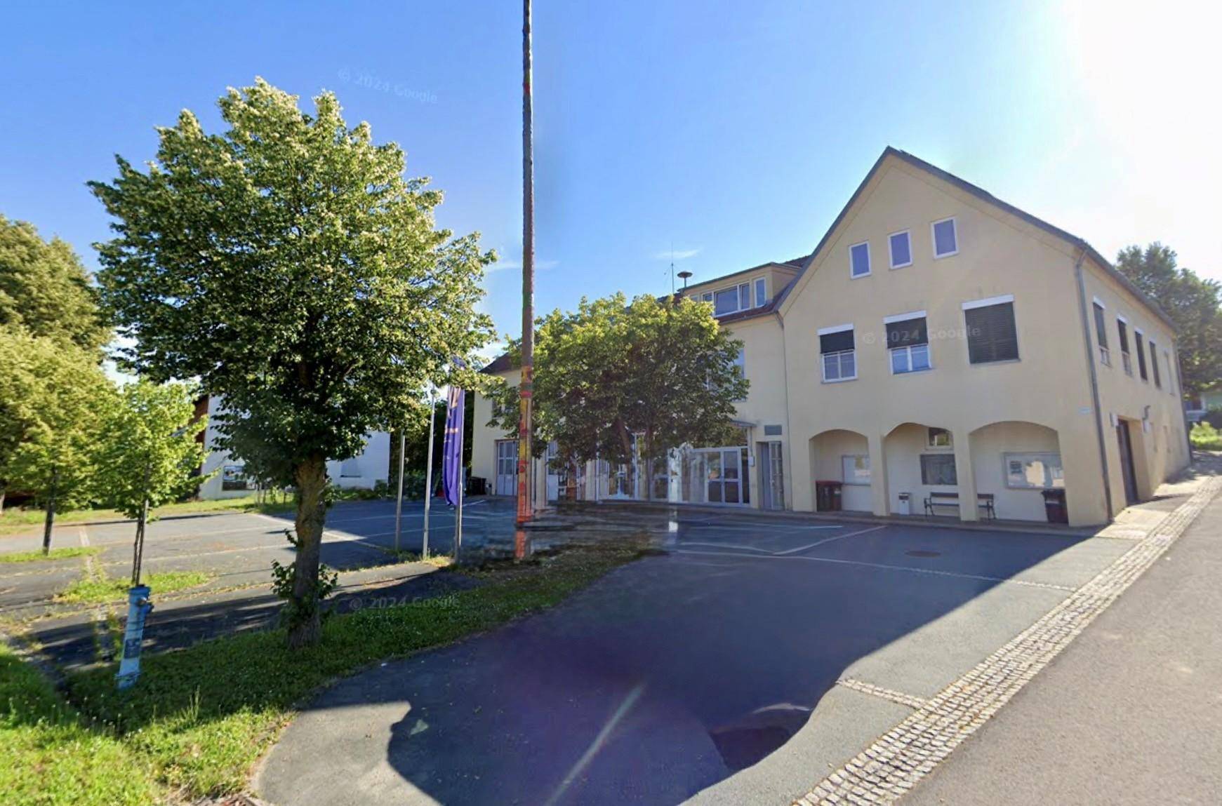 Ansicht des Gebäudes mit Eingangsbereich, Parkplätzen und Bäumen an einem sonnigen Tag.