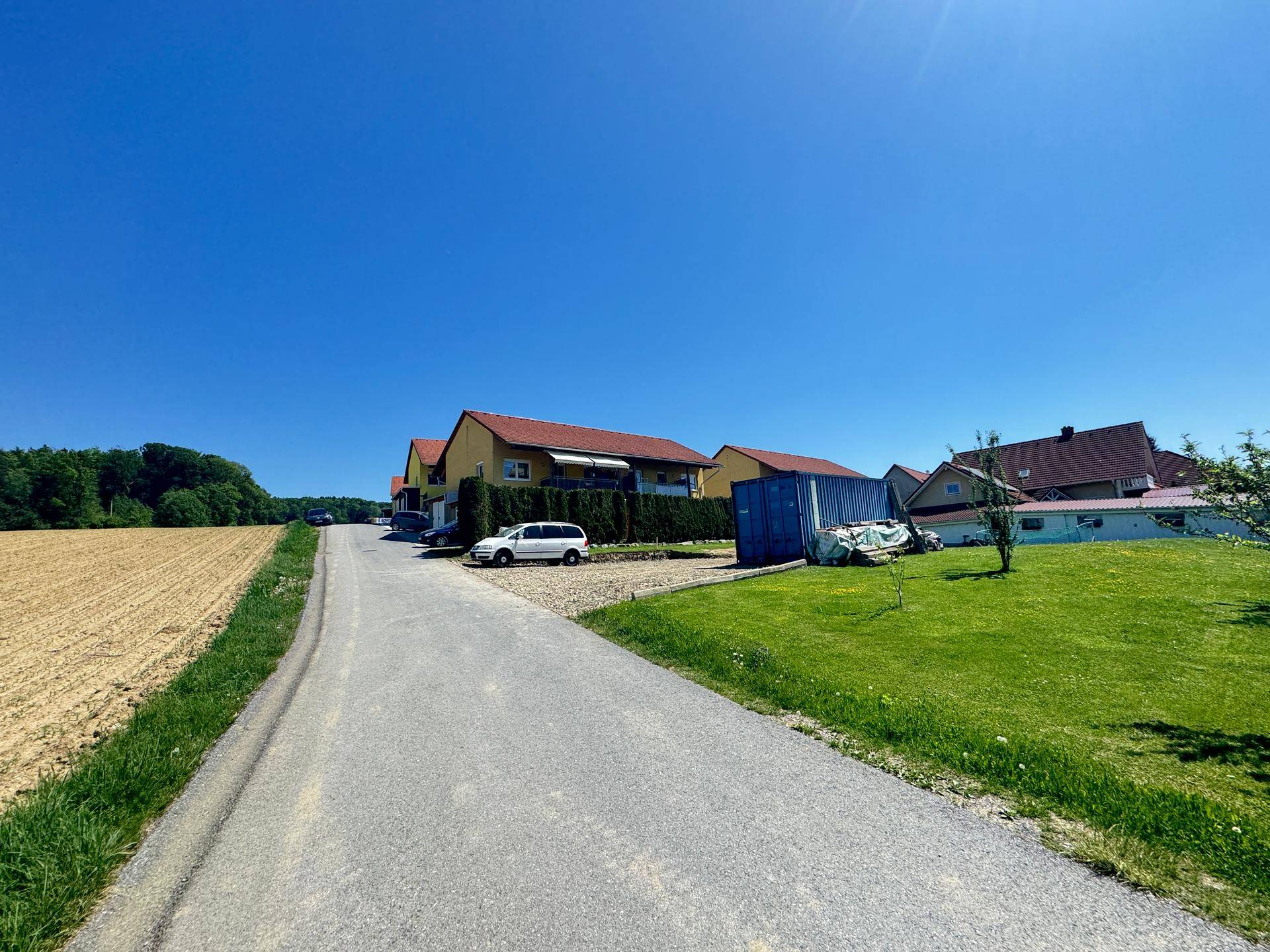 Ländliche Straße mit Feldern und Wohnhäusern in der Ferne unter strahlend blauem Himmel.