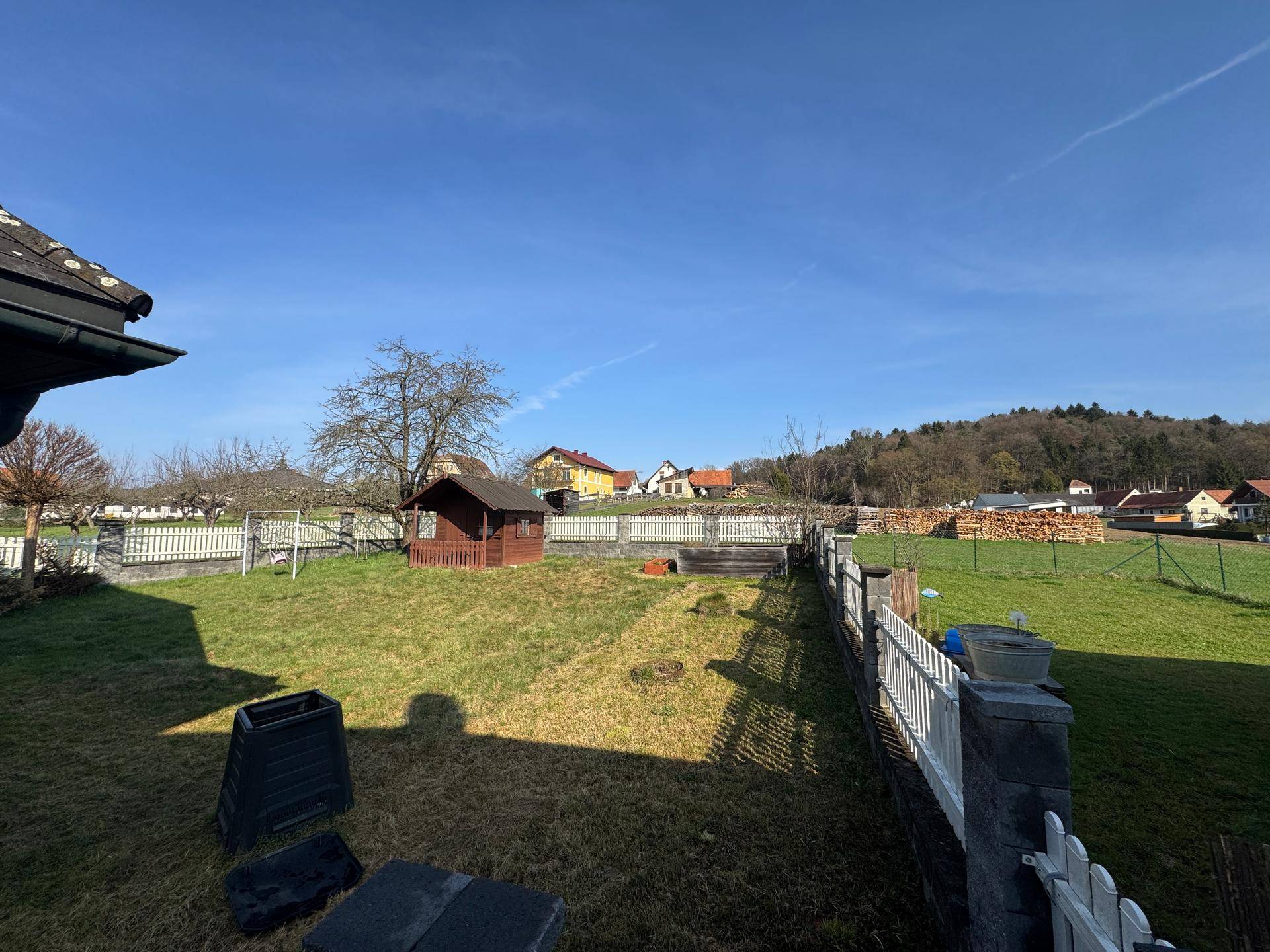 Großer Gartenbereich mit Rasenfläche, kleinem Holzschuppen und weißem Lattenzaun.