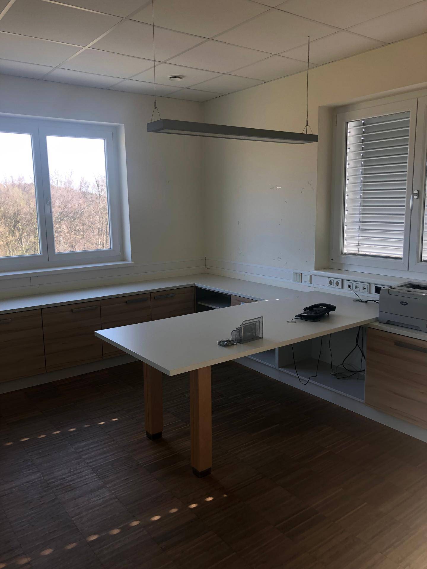 Heller Büroraum mit großem Schreibtisch, Holzschränken und Jalousien an den Fenstern.