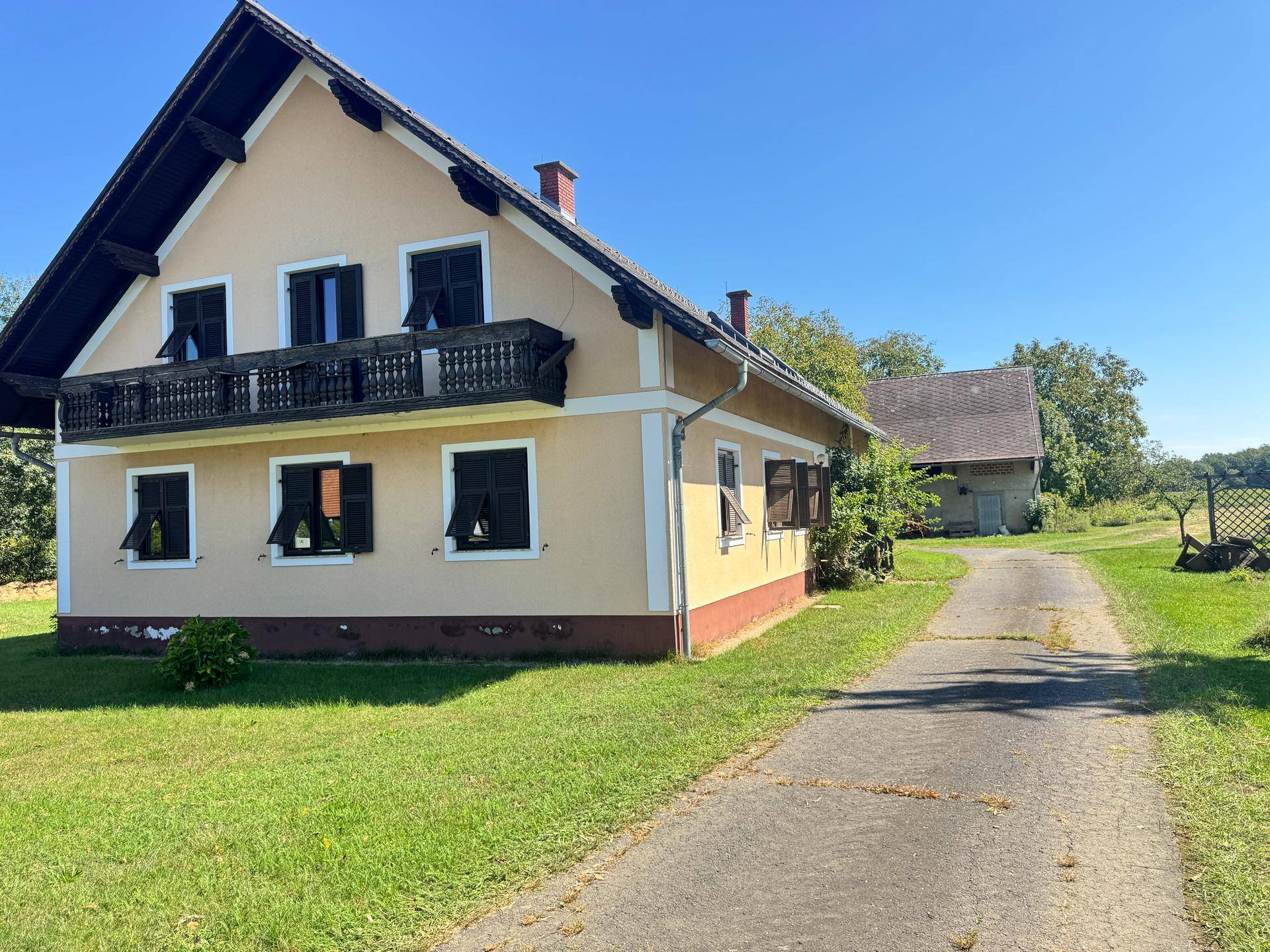Traditionelles Landhaus mit Balkon und Zufahrt, umgeben von grüner Landschaft.