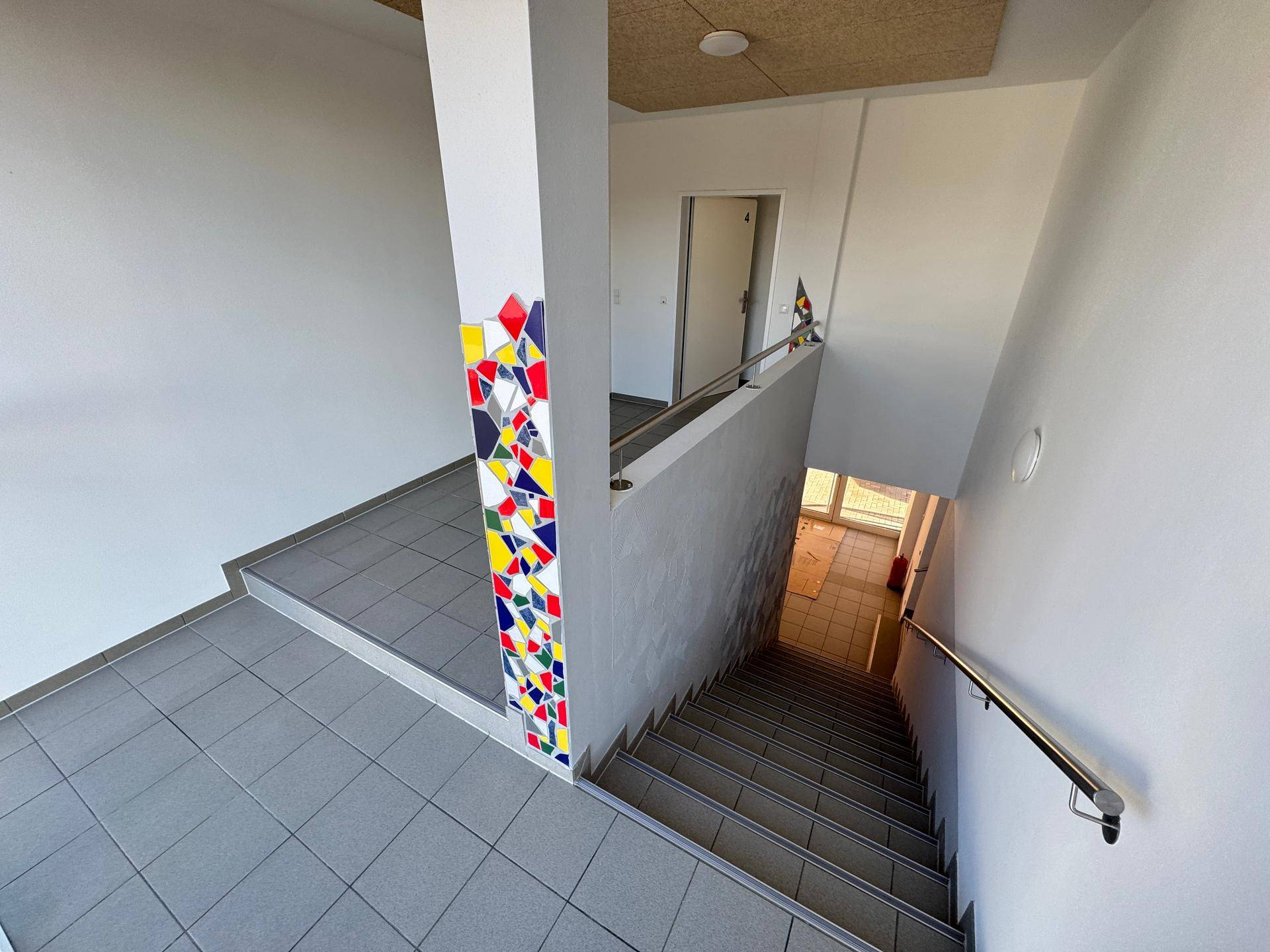 Treppenhaus mit farbiger Mosaikverzierung an der Wand und Fliesenboden.