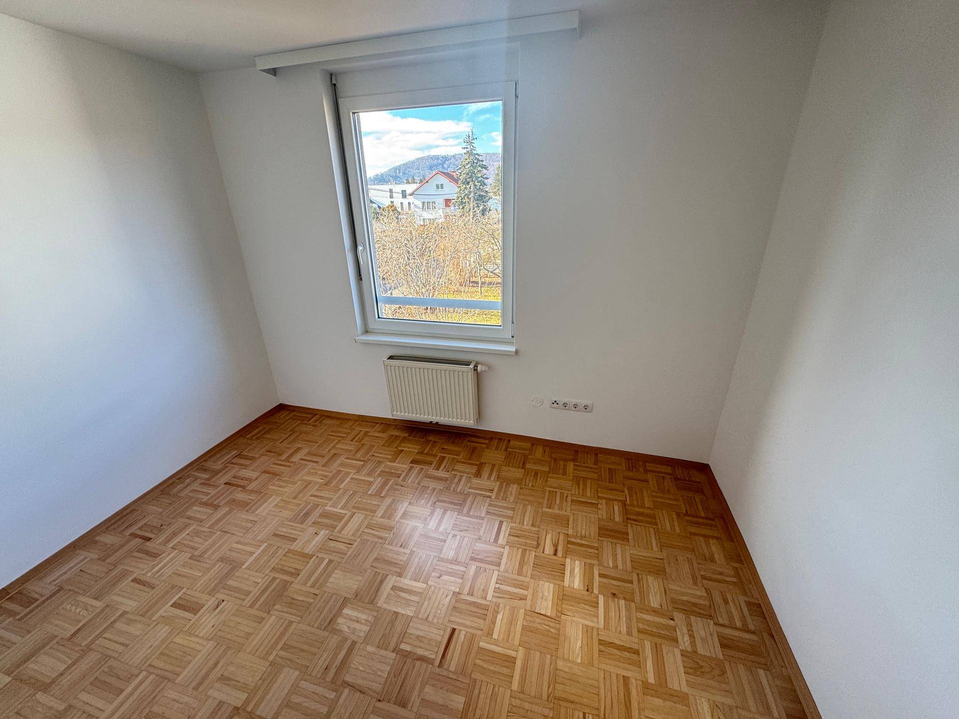 Heller Raum mit Parkettboden und großem Fenster mit Blick ins Grüne.