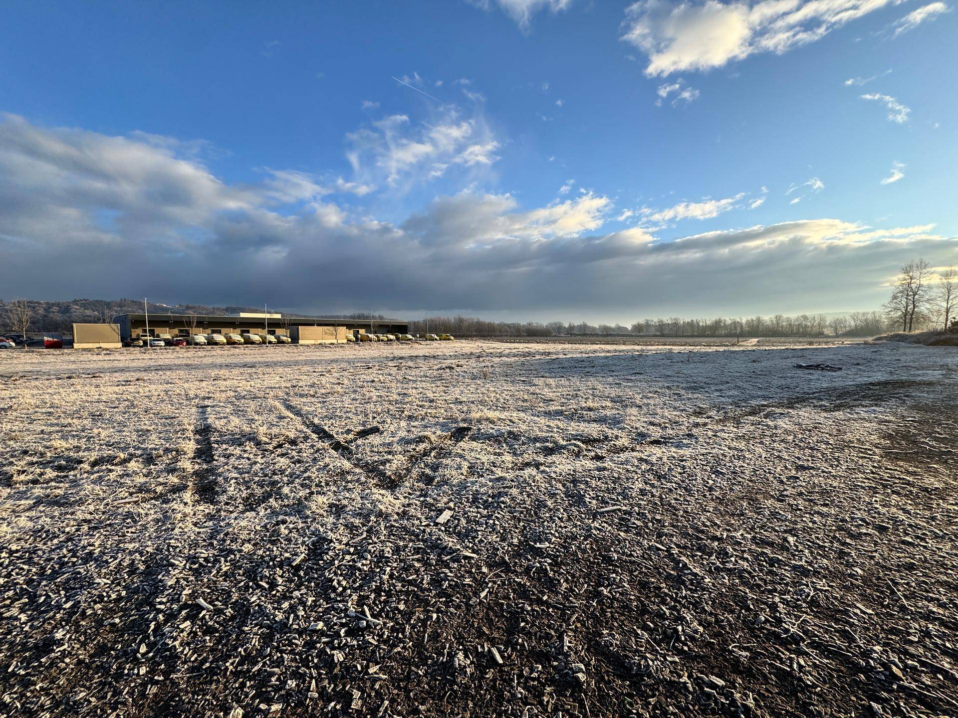 Weitläufiges, frostbedecktes Feld mit Gewerbegebäuden im Hintergrund unter einem teilweise bewölkten Himmel.