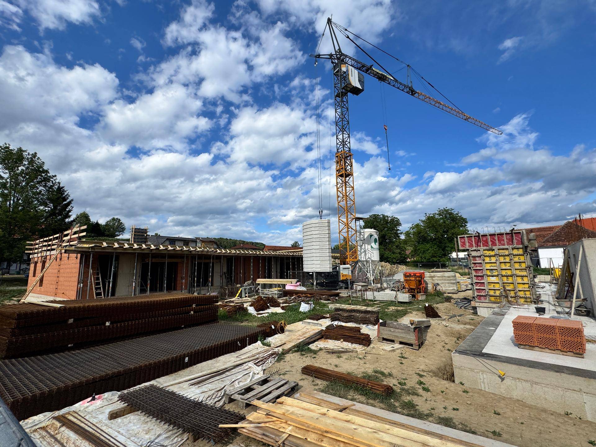 Weitläufige Baustelle mit Kran, Rohbau und Baumaterialien unter blauem Himmel mit Wolken.