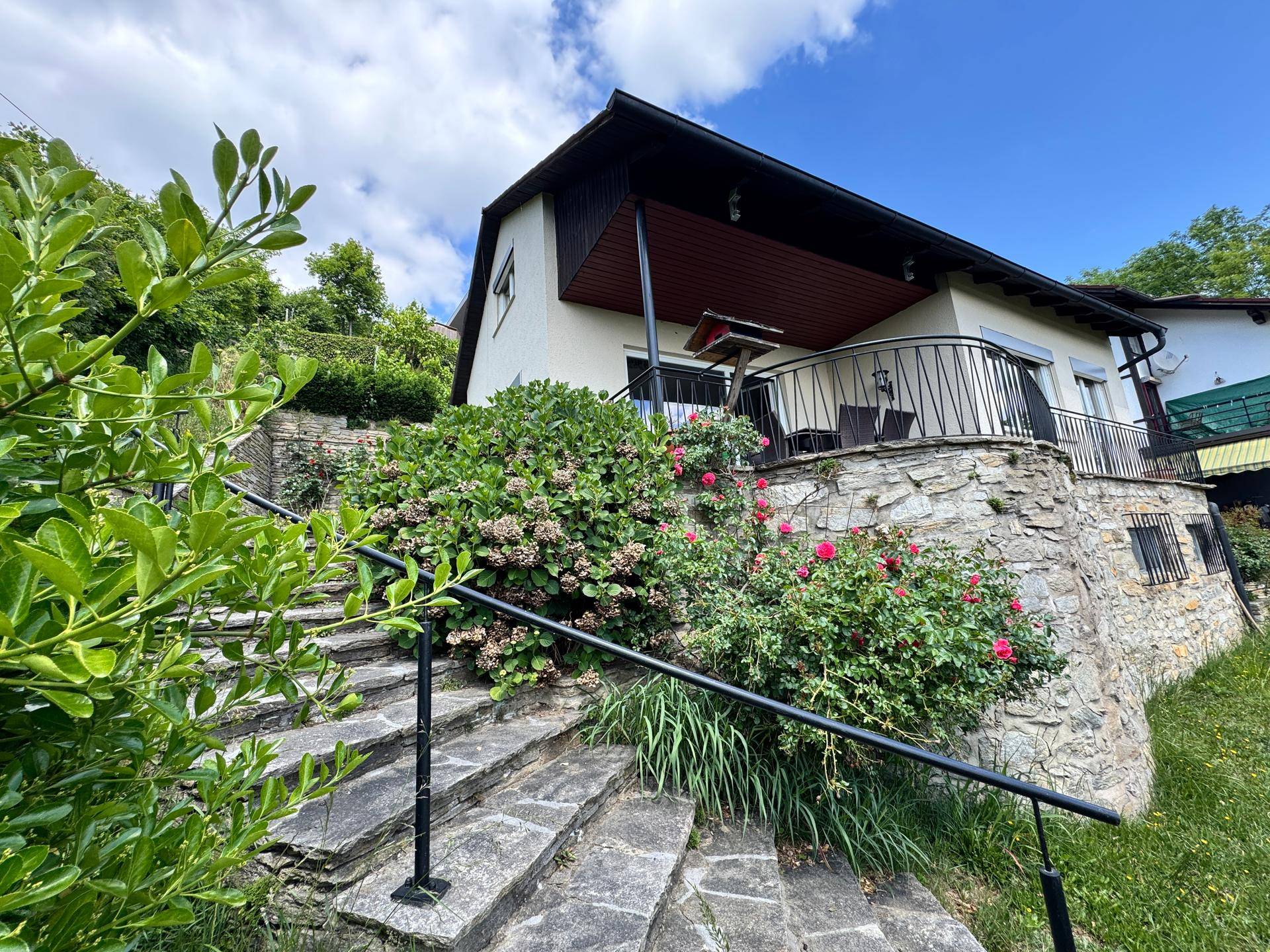 Seitlicher Blick auf das Haus mit Steintreppen, üppigen Büschen und einem Balkon mit schmiedeeisernem Geländer.