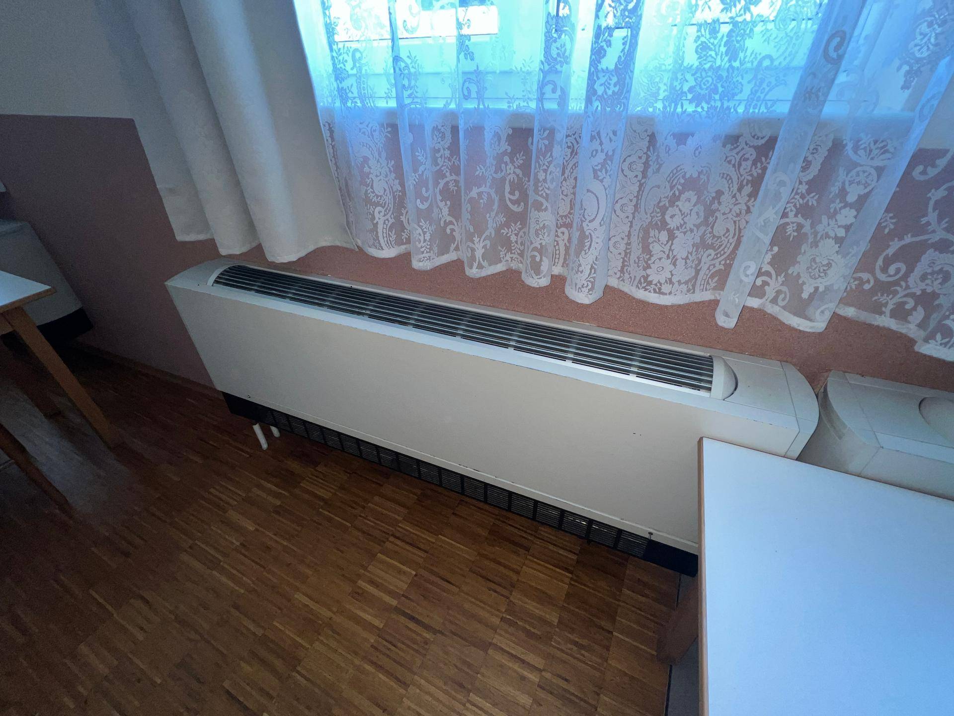 Einfaches Zimmer mit Holzfußboden, großem Fenster und Heizkörper unter der Fensterbank.
