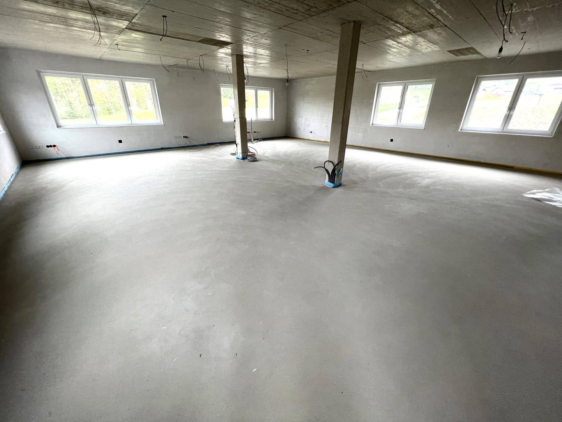 Großer, unfertiger Raum mit Betonboden und vielen Fenstern, bereit für den Innenausbau.